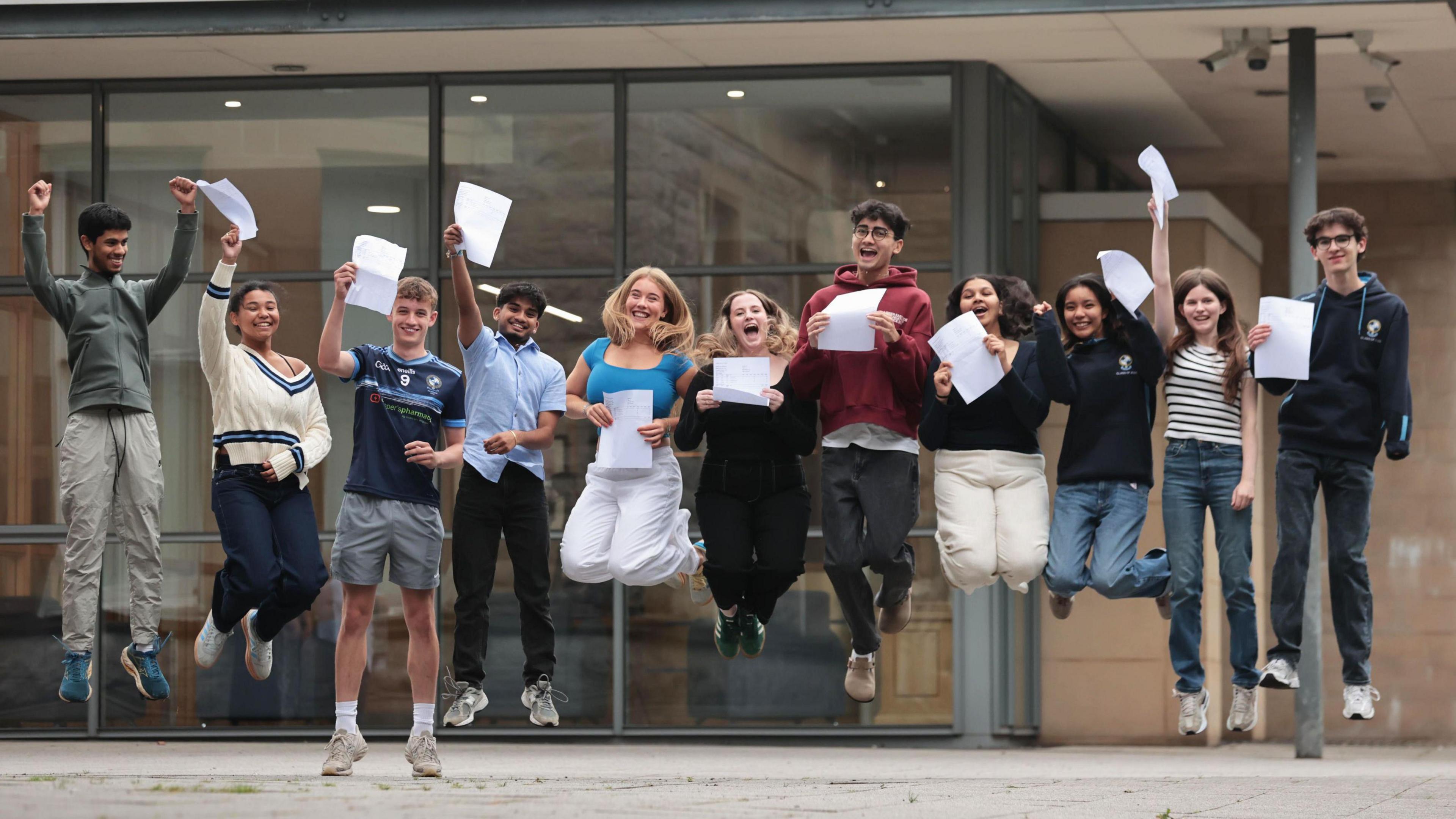 Students receiving their A-level results at Rathmore Grammar School in Finaghy, Belfast - 11 of them, a mix of boys and girls, all happily jumping in the air outside the school with their paper results in hand