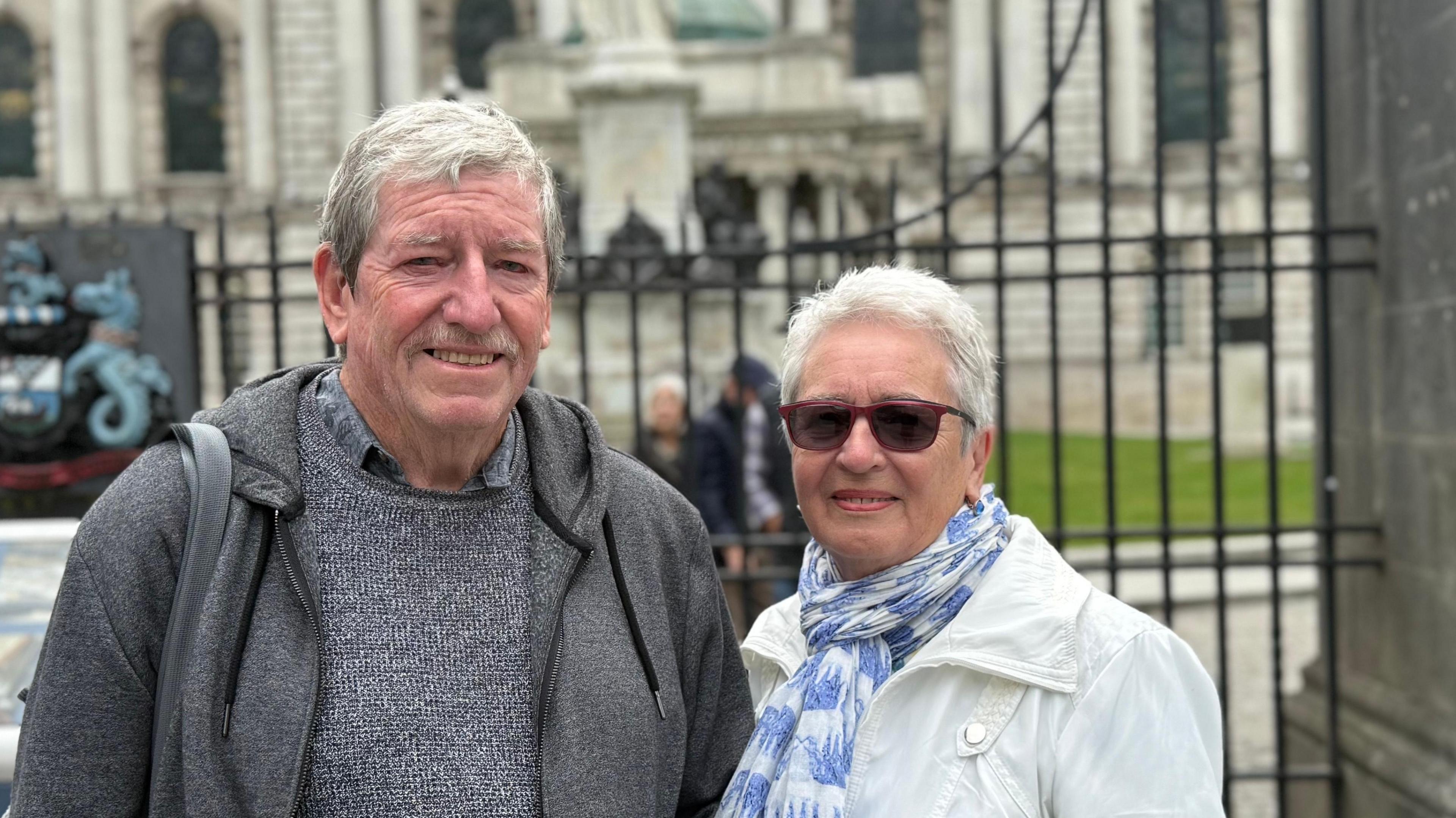 A man and a woman standing outside railings, behind them a grand looking white washed Belfast City Hall.
The man has grey hair swept to the side and blue eyes. He is smiling at the camera. He is wearing a grey hoodie over a grey jumper.
The woman is a head or so smaller than him and has white short hair. She is wearing a white coat with a white and blue scarf on.