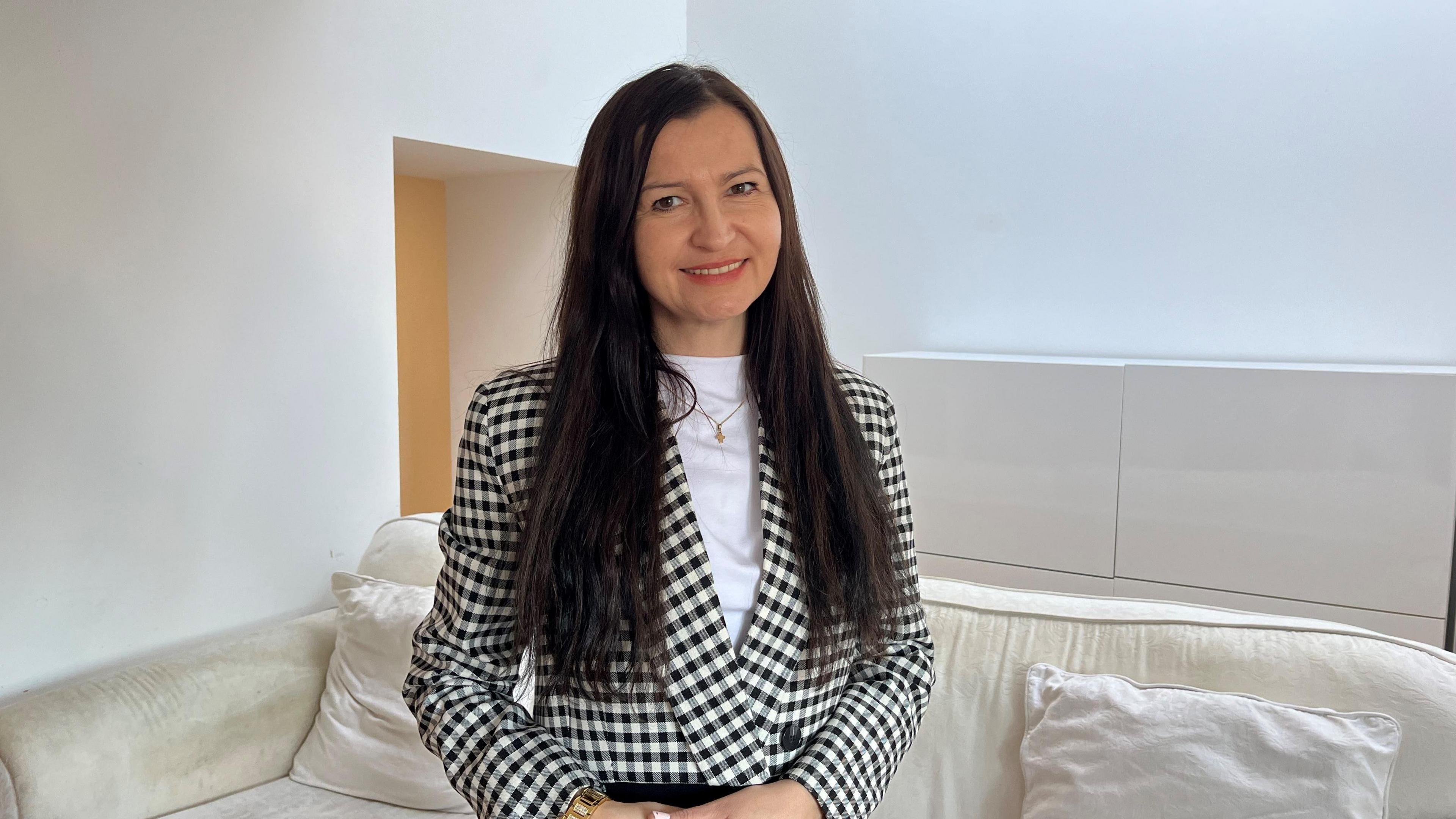 Helena Filipczak, who has long dark hair and brown eyes, smiles at the camera while standing in front of a sofa in a white room. She is wearing a white top and a black and white checked jacket.