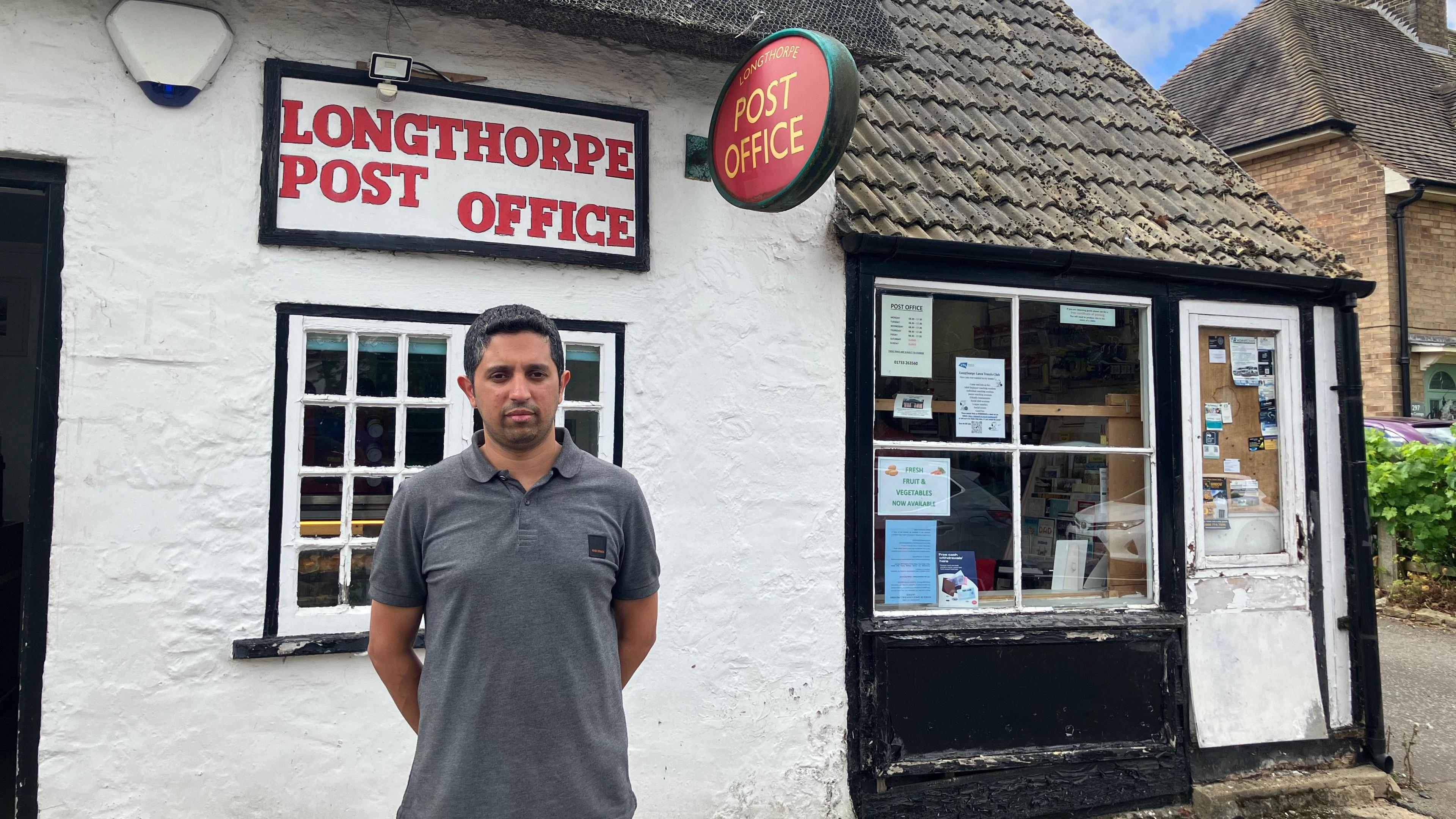 Arif in a grey T-shirt standing outside a white stone building with a sing that reads "Longthorpe Post Office"