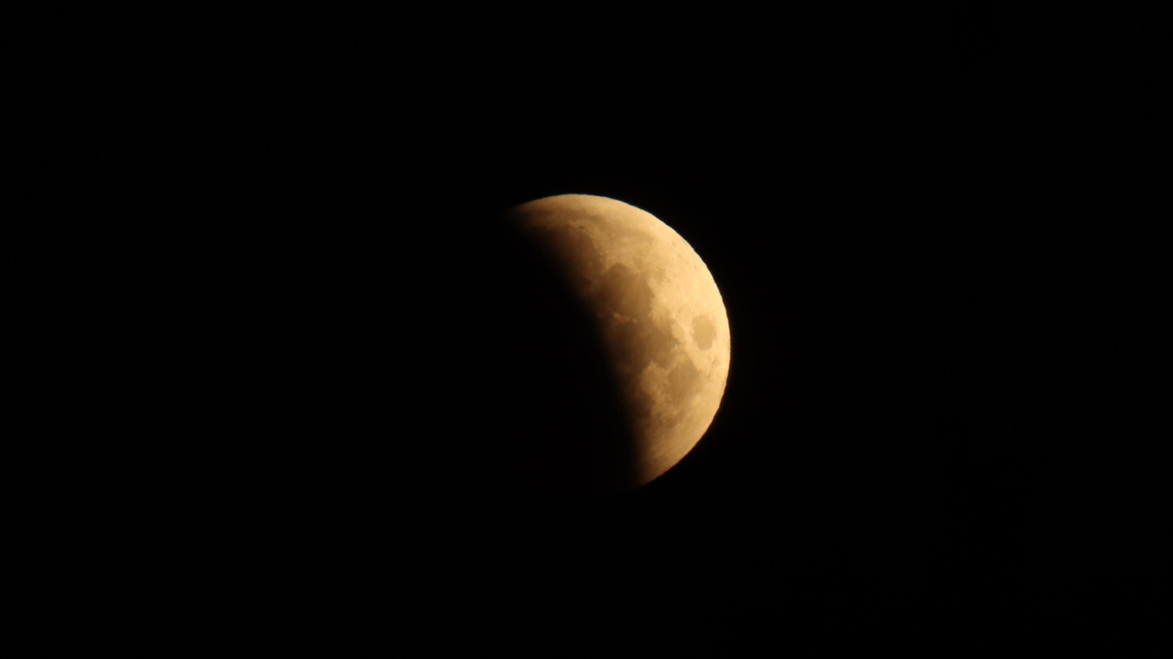 The moon surrounded by darkness. It is partially obscured by a lunar eclipse. It is orange in colour. 