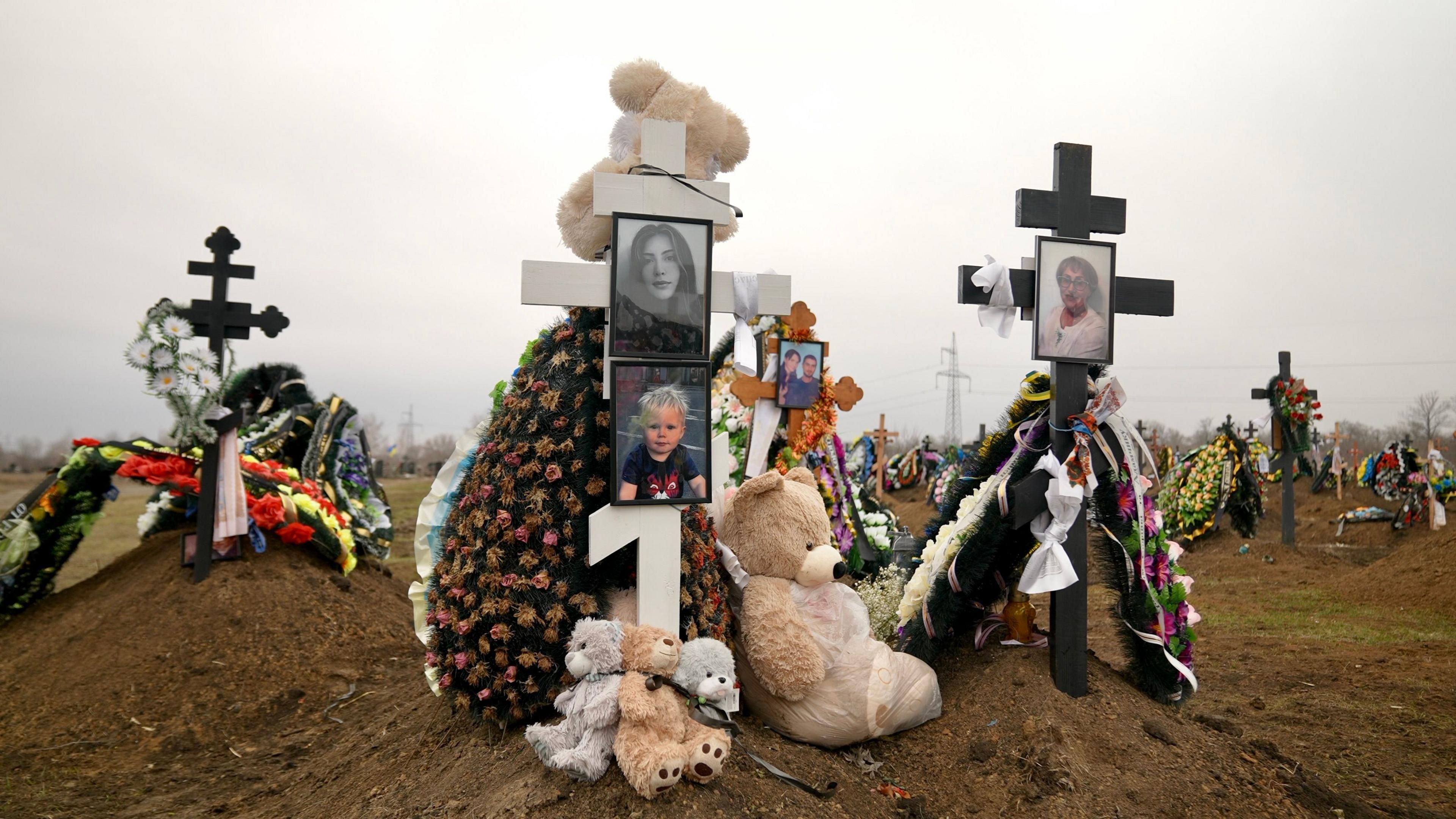 A number of wooden crosses within a cemetery. One on the right is the grave of Tetiana Tarasevych and displays her photos. The cross on the left is the grave of Sophiia Buhayova and her son Adam. Both graves are covered in artificial flowers and teddy bears. 
