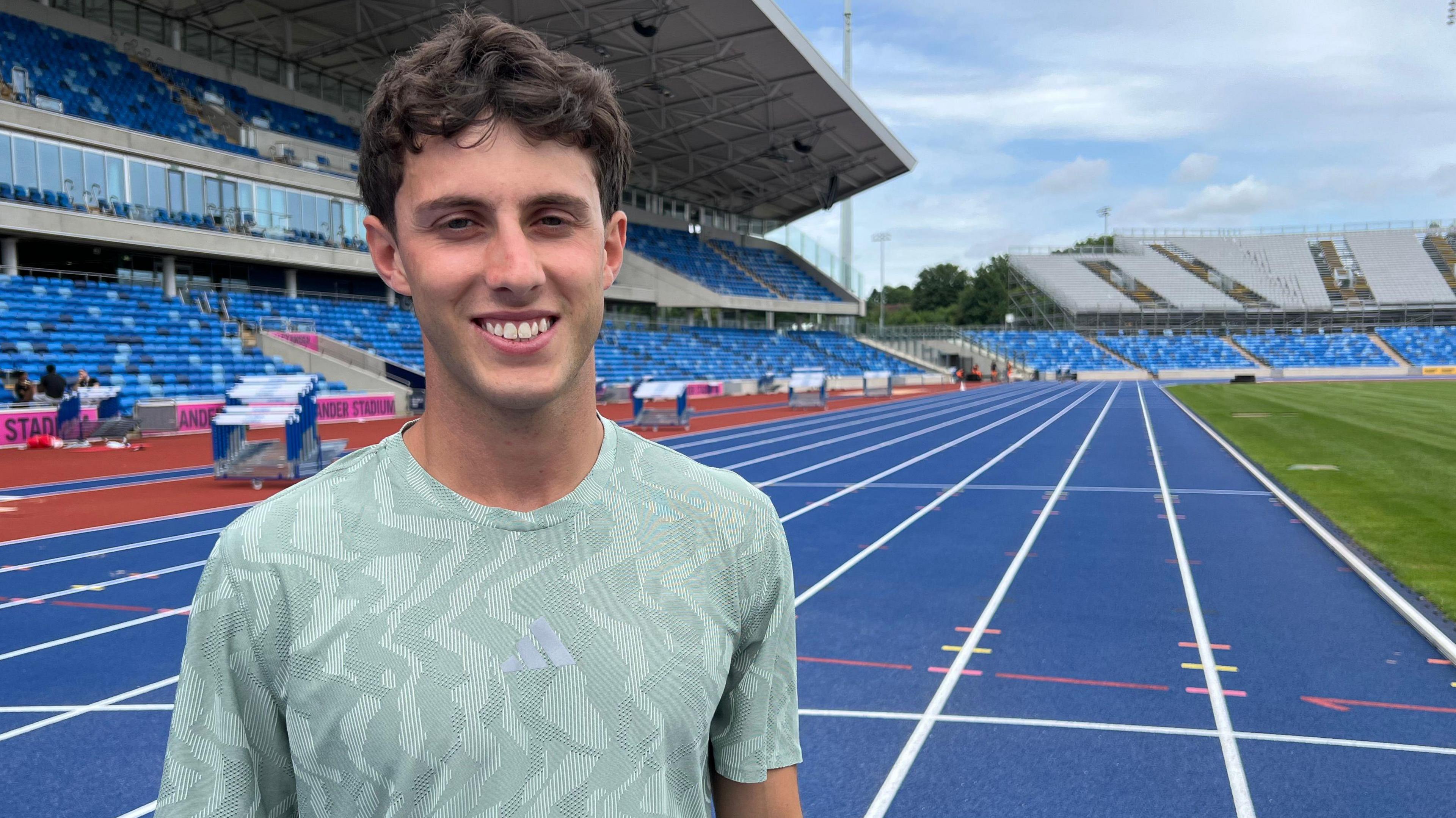 Jake Minshull stands in a light green training top on a blue race track. The white lane lines stretch into the background. The stands are behind him and the grass infield on the right 