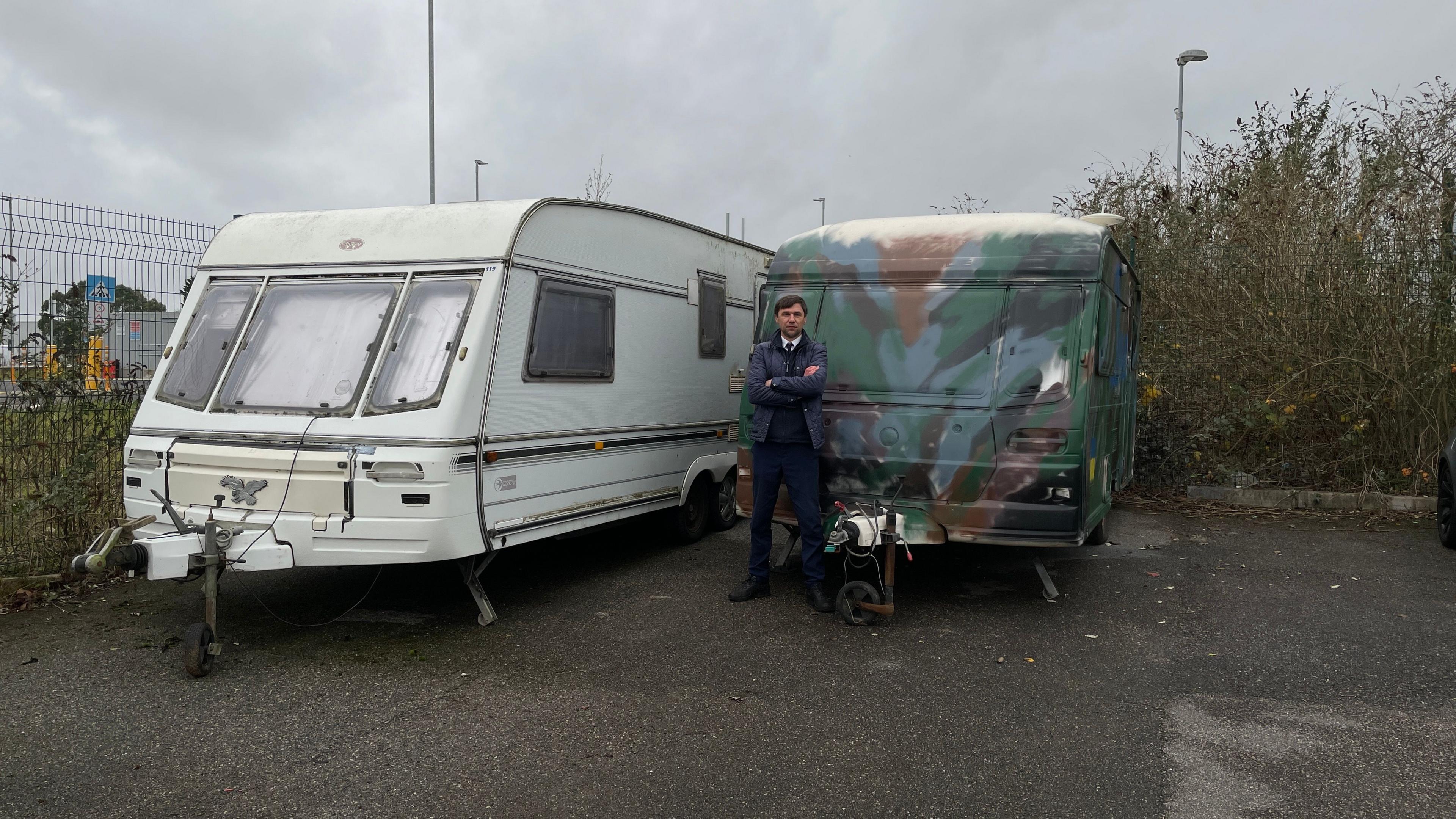 Maksym stands in front of two caravans. One is painted in Ukrainian army camo.