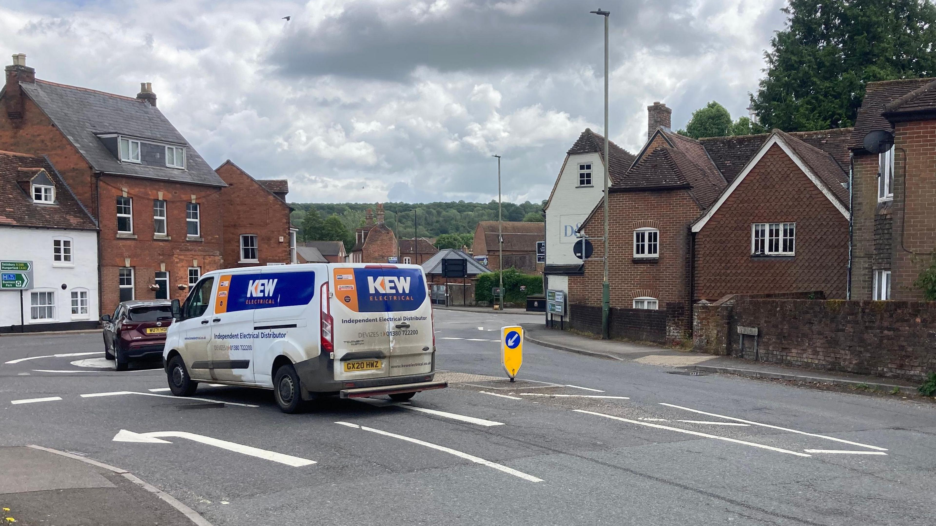 New Road in Marlborough. There is a mini roundabout and a van and black car travelling on it. Buildings can be seen along the road.