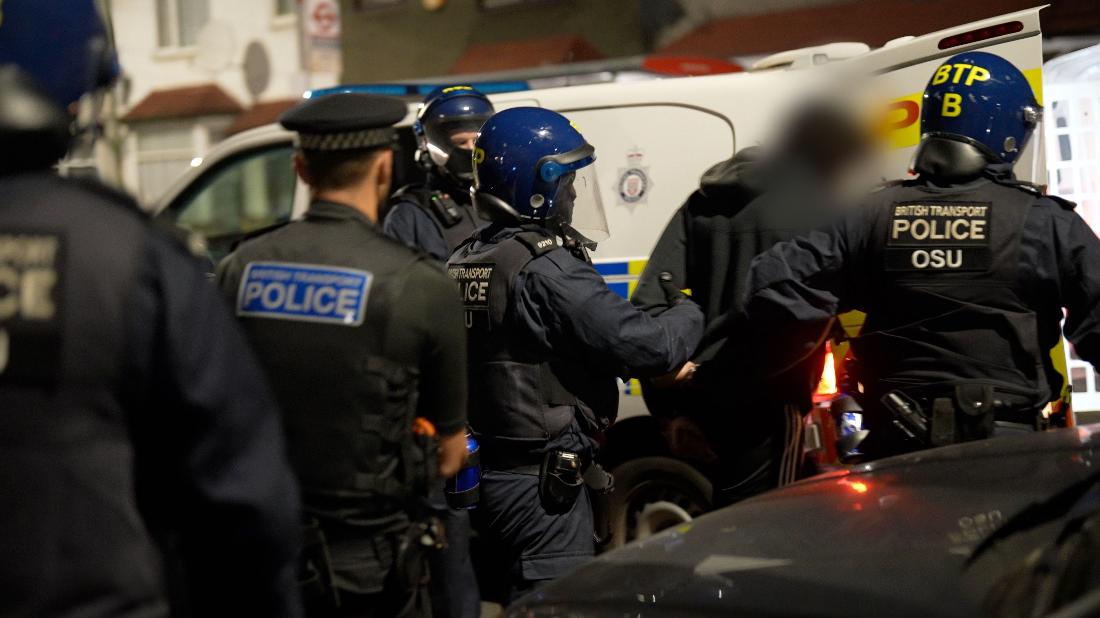 Two British Transport Police officers lead a robbery suspect into the back of a police van, they are wearing helmets and body armer. Two other police officers follow them, with another stood by the van.