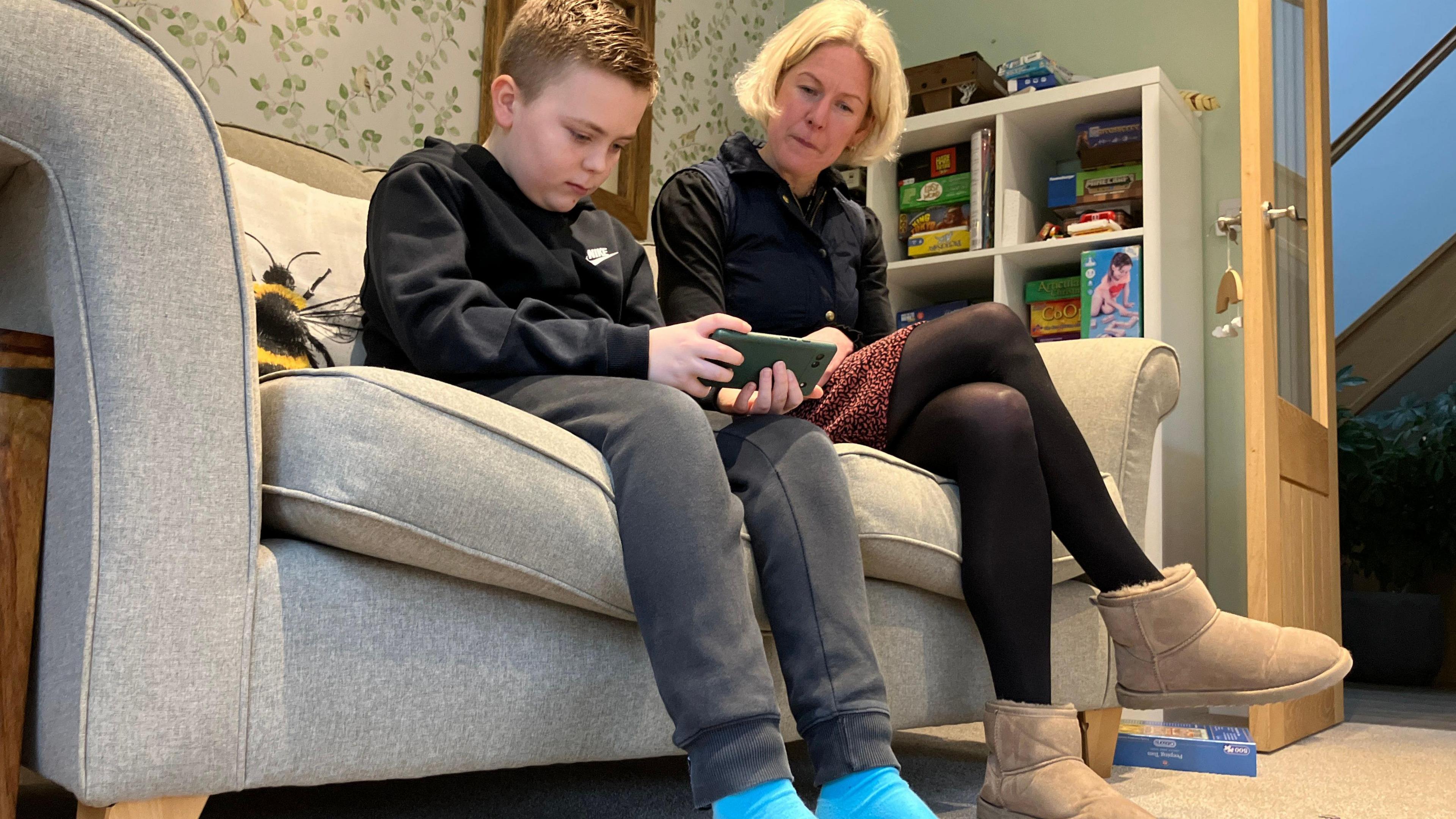 A young boy in a black top looks down at a phone while a faired haired woman looks at him.