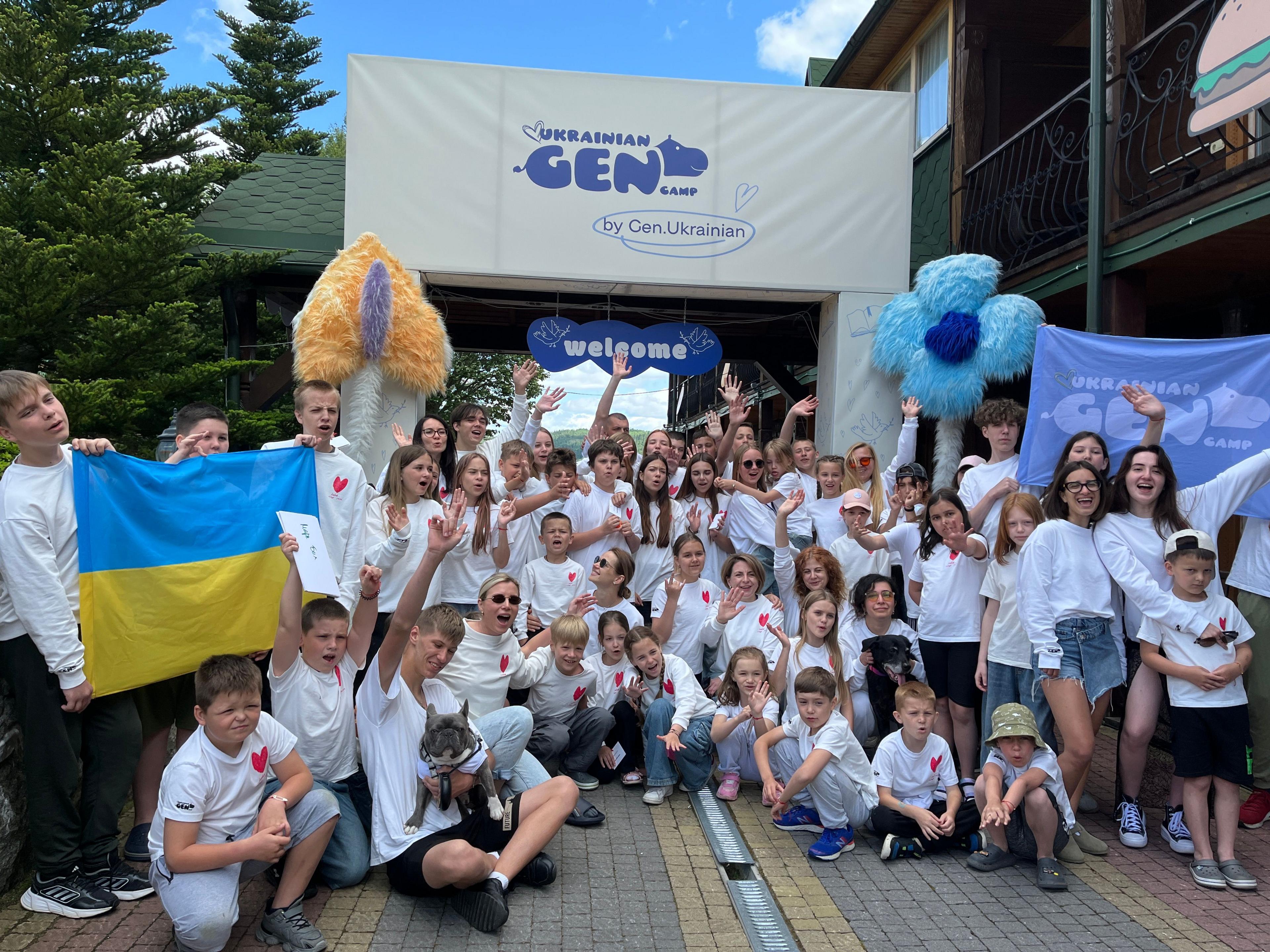 A group of children, wearing matching white tops with a red heart logo, pose for a group photograph, waving their hands, smiling and holding Ukraine flags.