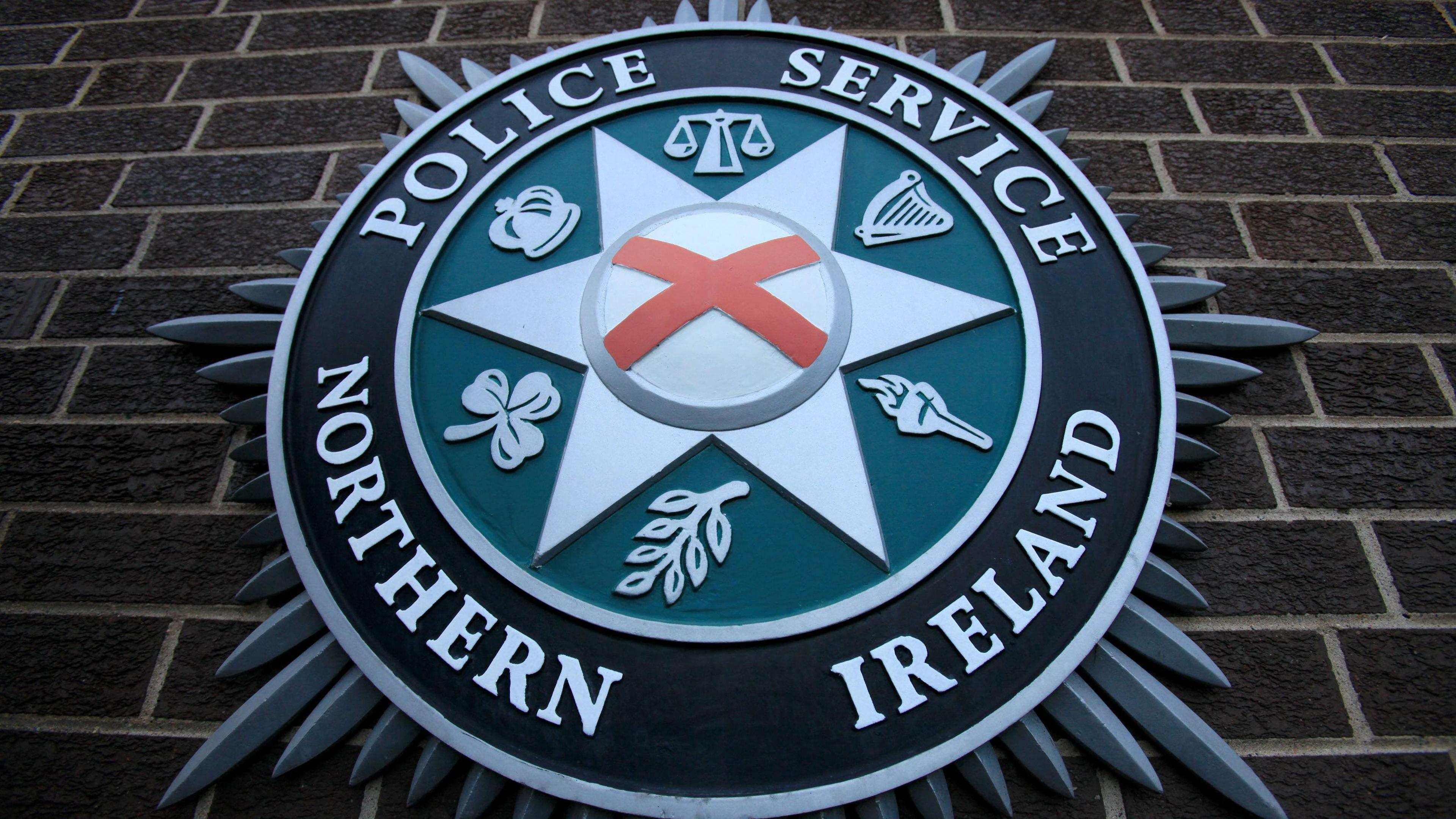 A close-up of a Police Service of Northern Ireland crest on the brown-bricked wall of its Belfast headquarters. The centre of the crest shows a red cross on a white background, which is inside a white star, inside a green circle, which is inside a larger black circle, all surrounded by silver spikes. There are six symbols in between the points of the central star, including a harp; a crown; a shamrock; a flaming torch; the scales of justice and an olive branch.