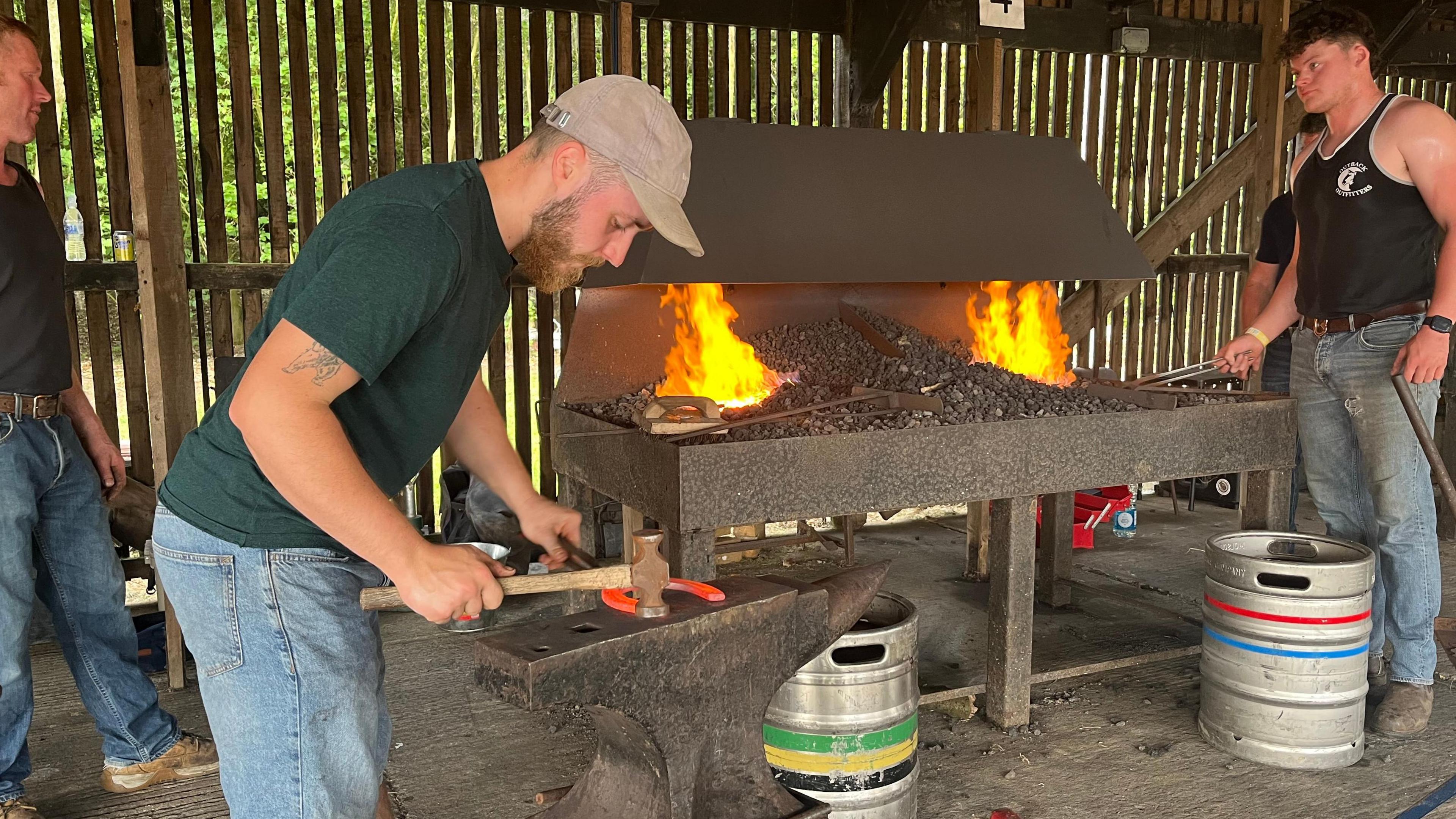 Two men pictured in front of a forge - one is using a hammer to hit a horse shoe. The other is looking towards the flaming forge.