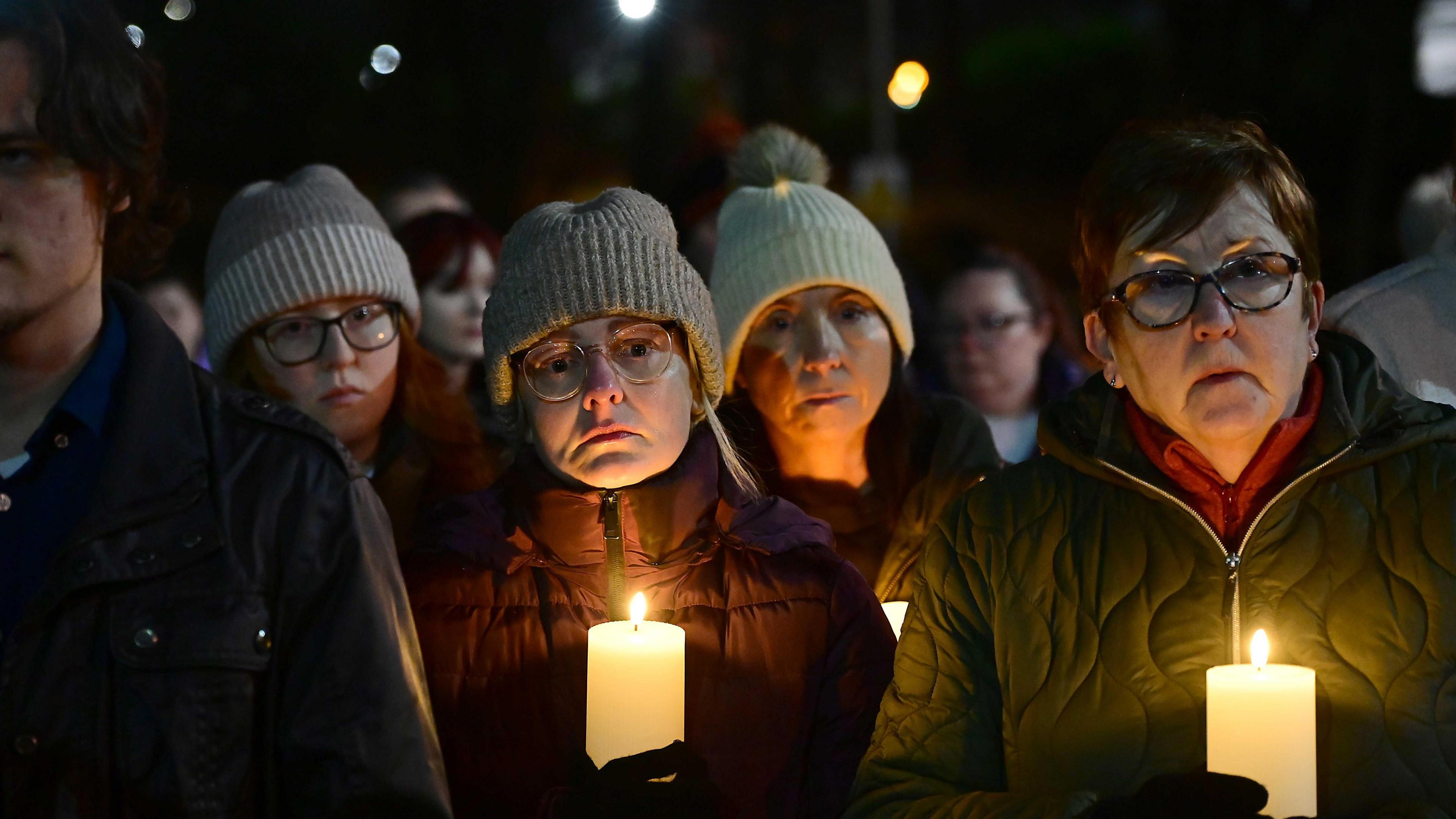 A close-up of four women and one man, who is partially obscured at the vigil. Three of the women are wearing woolen hats and three are also wearing glasses. Two of them are holding large white candles