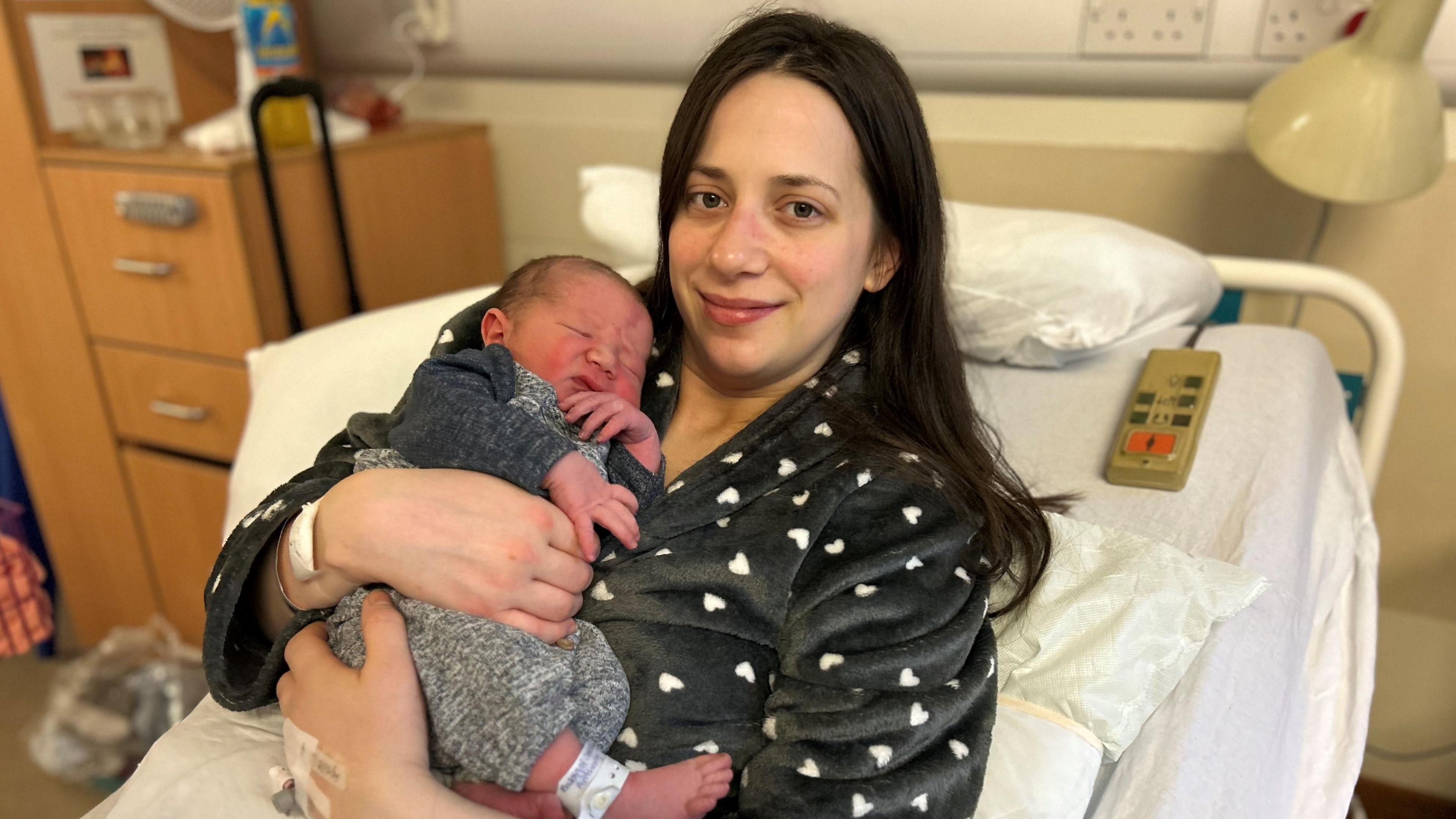 A baby in a grew outfit is held by a woman with long dark hair wearing black pyjamas with a heart motif