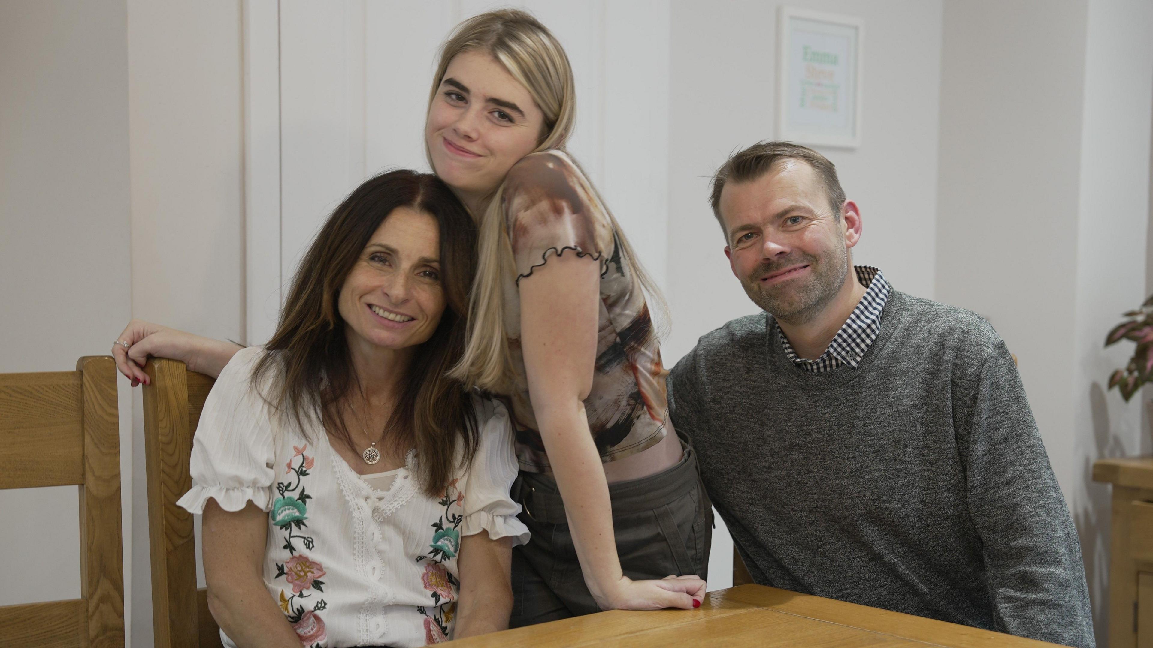 A teenage girl with long blonde hair stands between her mum and dad seated at the kitchen table. All three smile and look towards the camera.