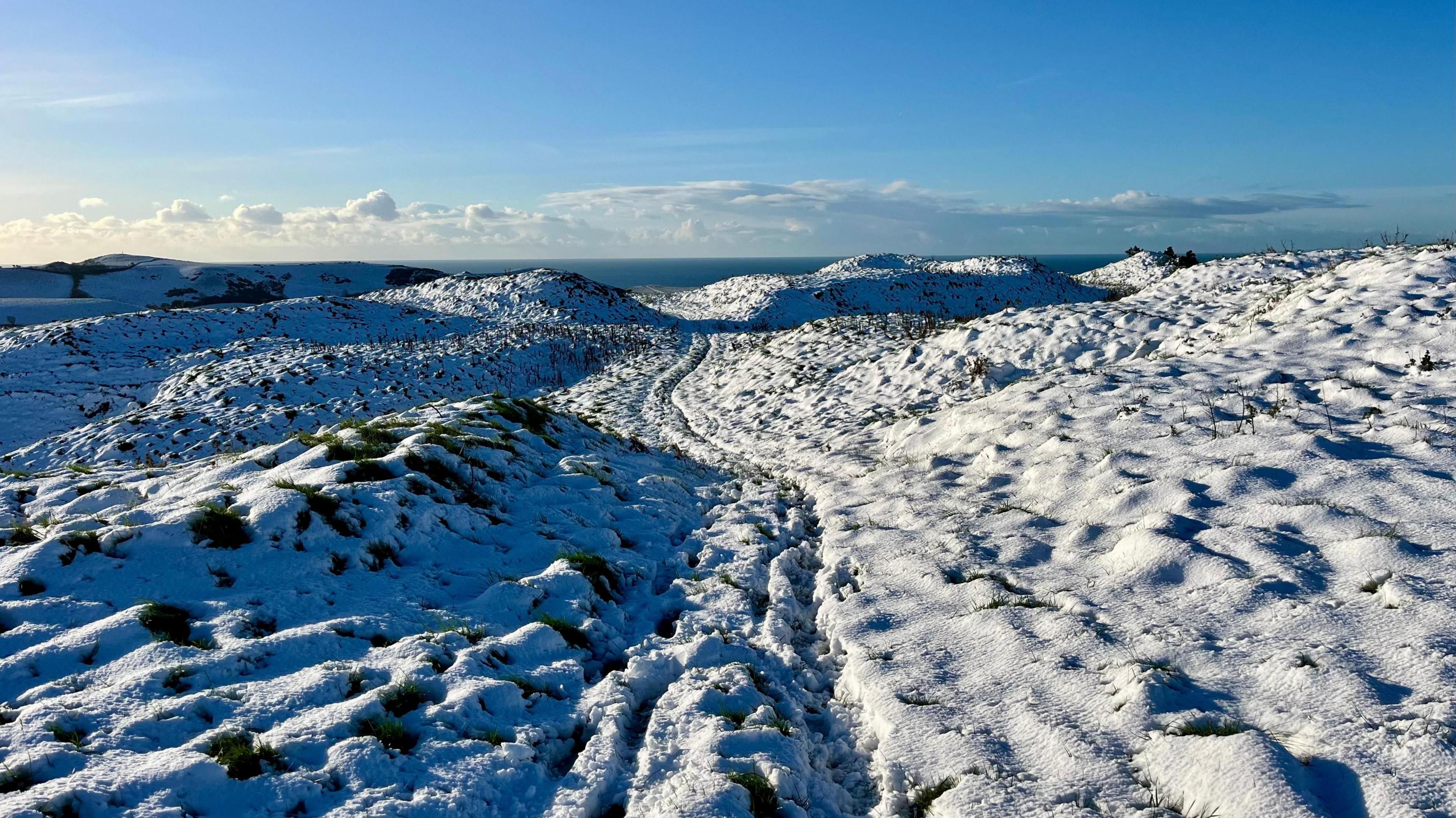 A landscape of hills covered in snow. The layer of snow is fairly thin with parts of the ground visible in places. The landscape is empty and bare under a fairly clear blue sky with just a few white clouds in the distance.