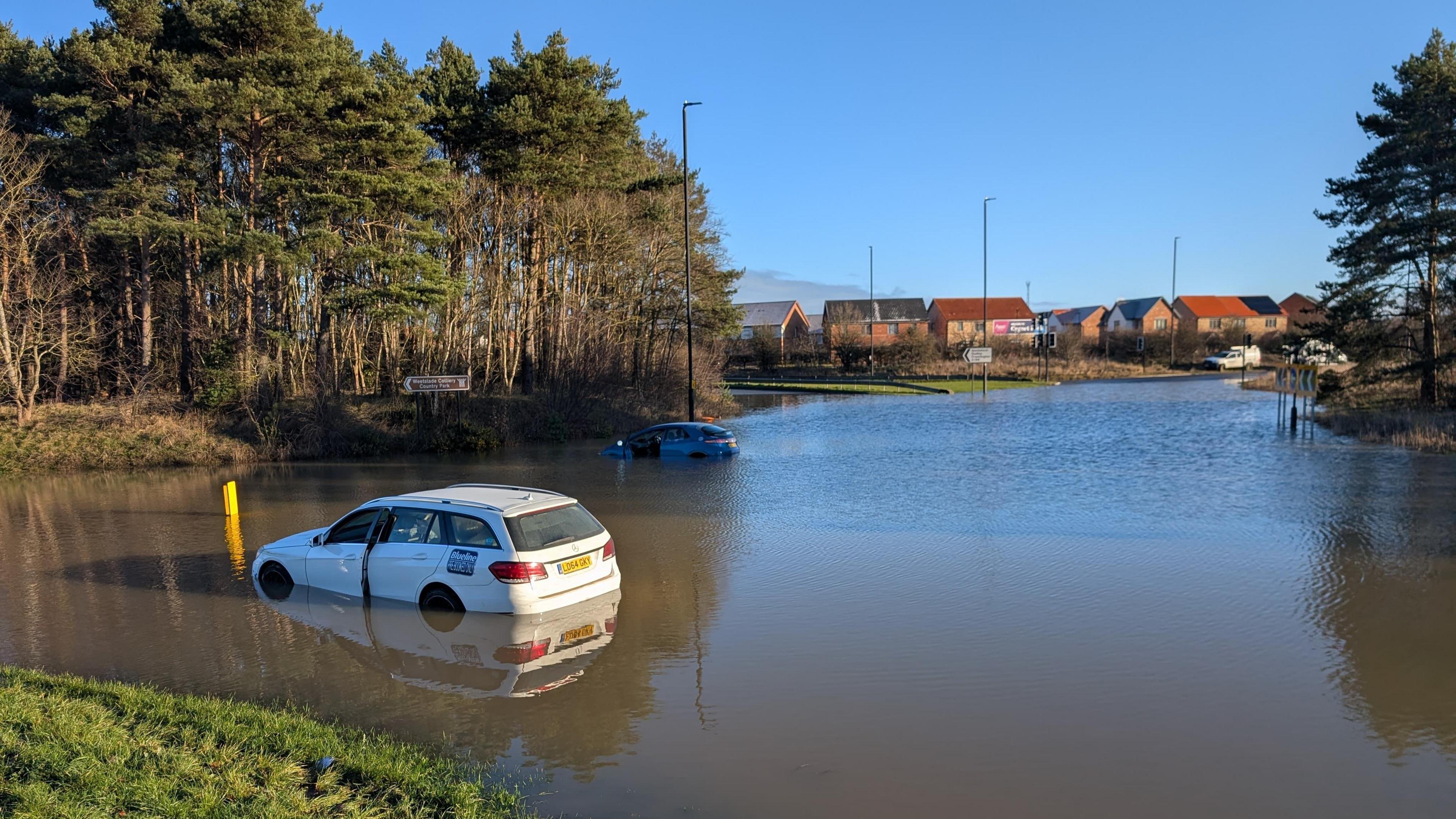 Sandy Lane Roundabout during the floods. Water covers the full three lanes of the roundabout. Two cars are partially submerged and stuck in the road. The water has covered part of the central roundabout and the roads leading from it. A housing estate can be seen in the distance.