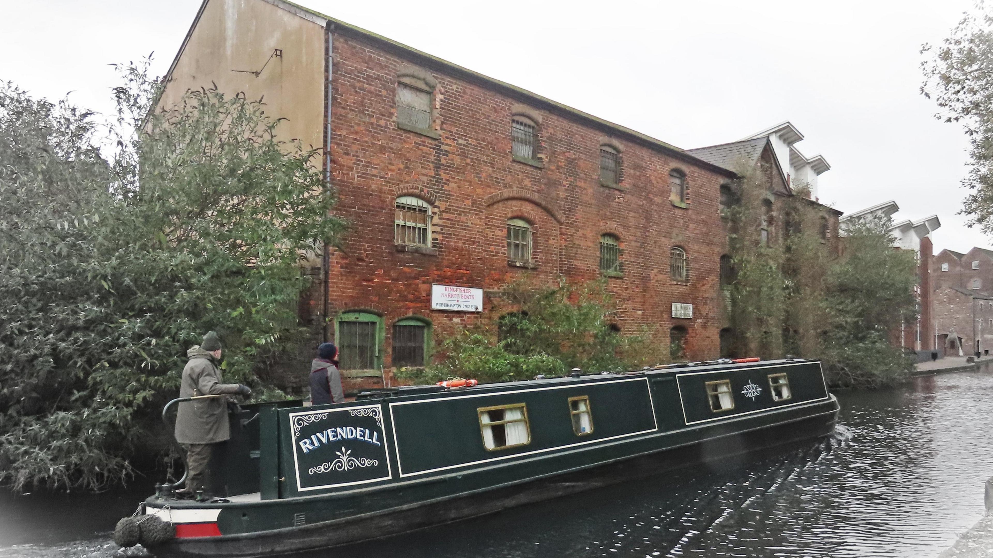 Two men are wearing dark coats and standing on a green narrowboat that says 'Rivendell'. They are driving along the canal with a red bricked building in the background.