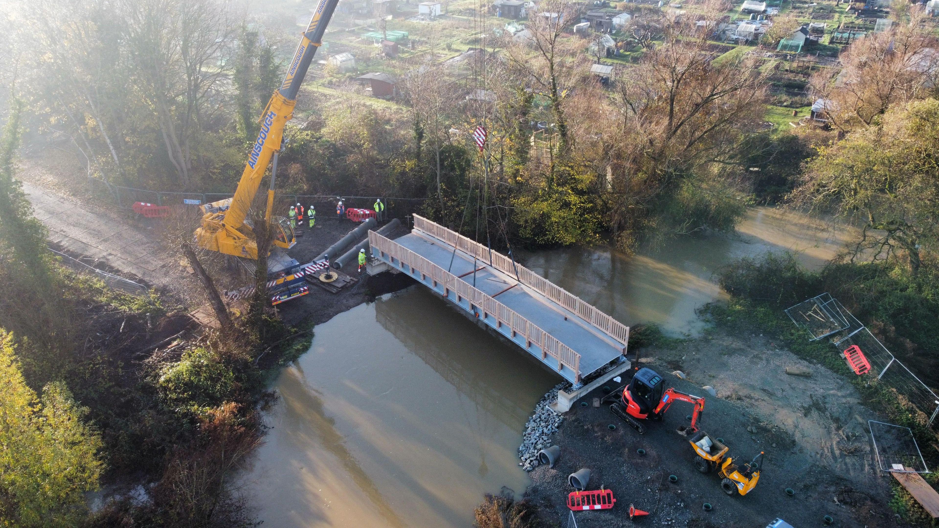 An aerial view of a footbridge over a river being hoisted into place by a yellow crane. Six people in high-vis yellow and orange jackets and hard hats stand on the riverbank near the crane. On the opposite bank a small red digger can be seen and a yellow vehicle used to transport waste. Trees line the riverbank.