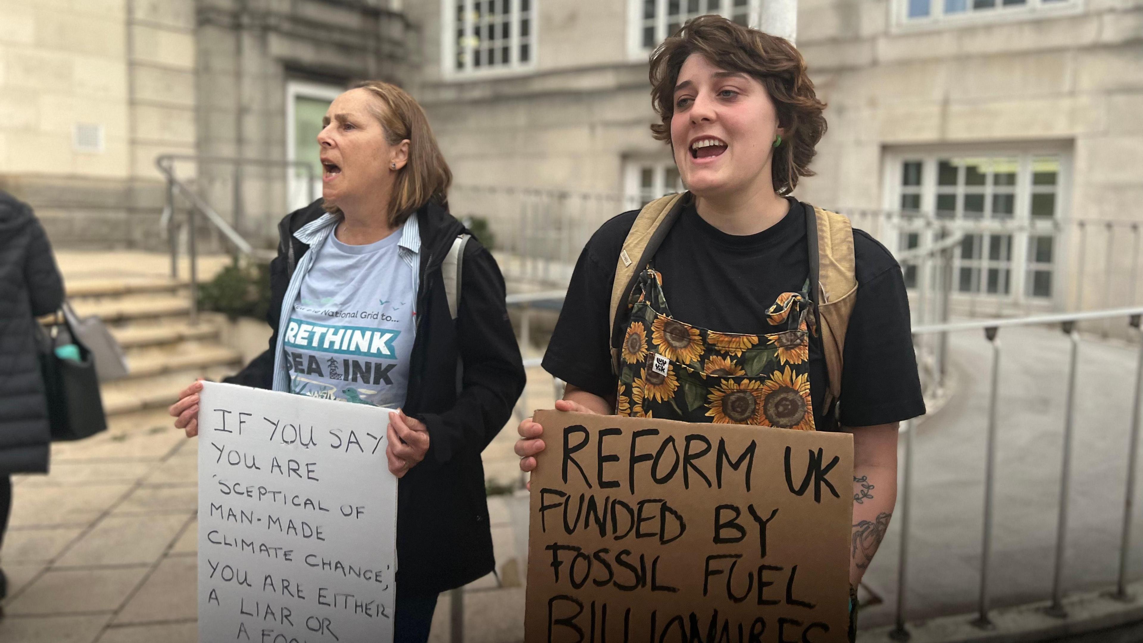 Two women stand outside Kent County Council's offices in Maidstone holding placards. One says "if you say you are sceptical of man-made climate change you are either a  liar or a fool and certainly a coward", the other says "Reform UK funded by fossil fuel billionaires." 