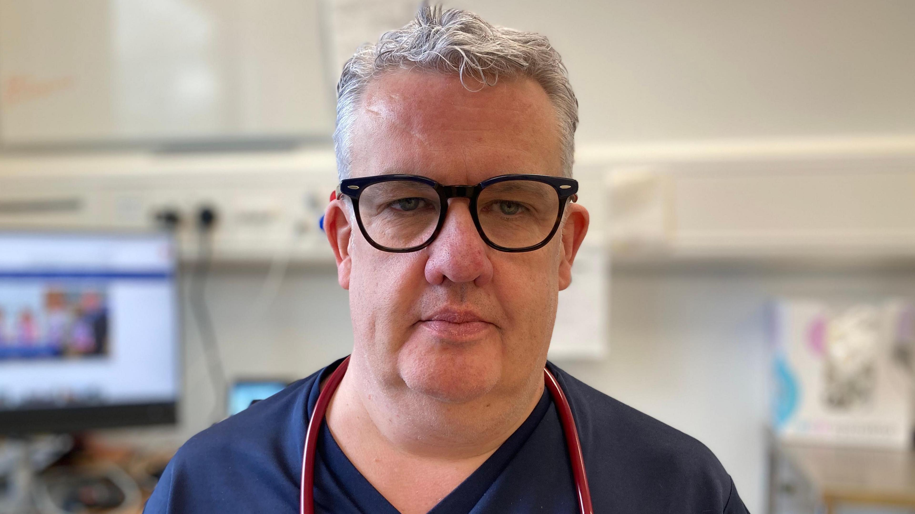 Dr McShane is wearing dark blue scrubs and black thick glasses, a red stethoscope is hanging over his shoulders. In soft focus in the background is a compute screen and a white board. 