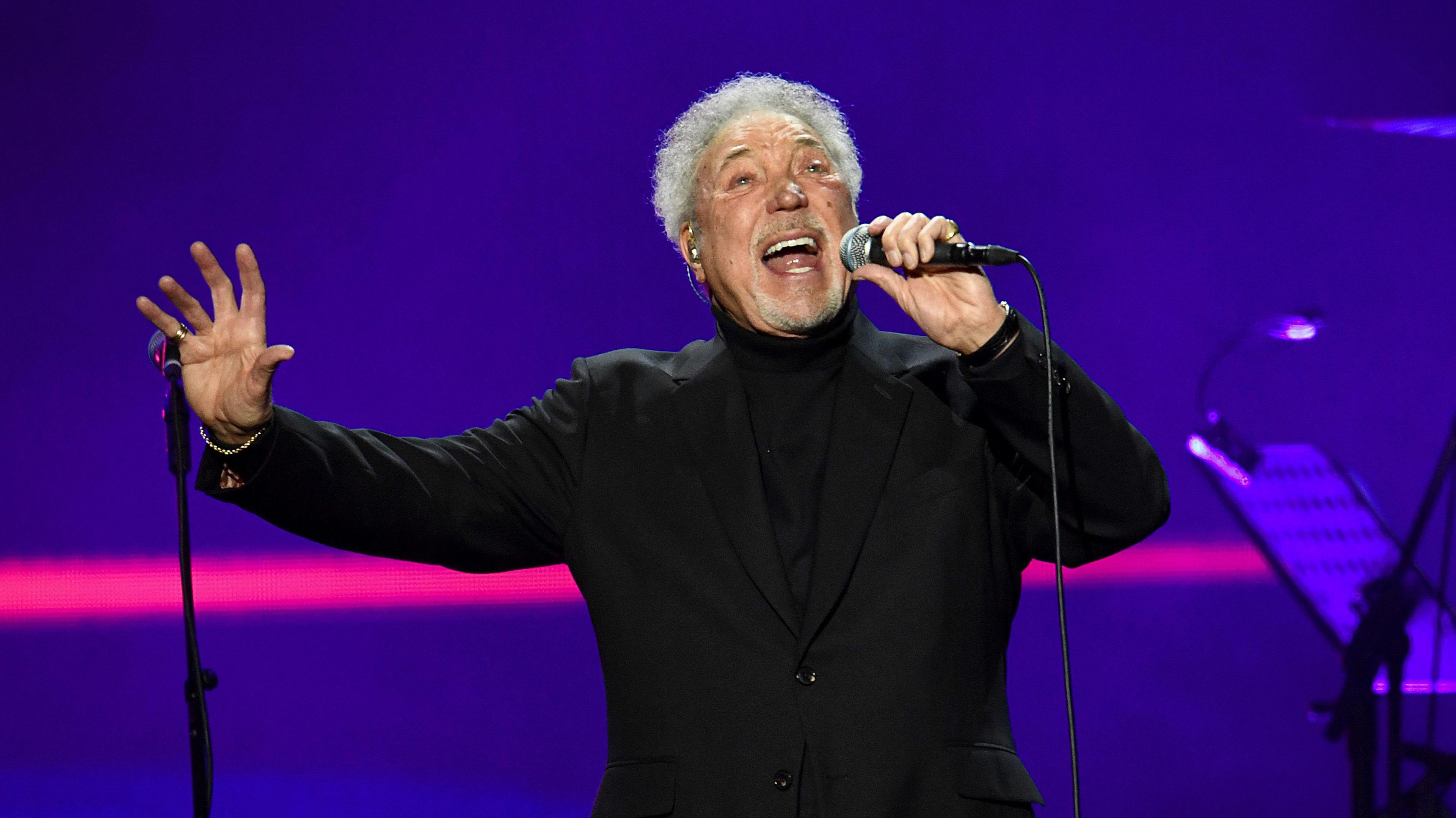 Tom jones holds a microphones in one hand and holds his other hand out as he sings 