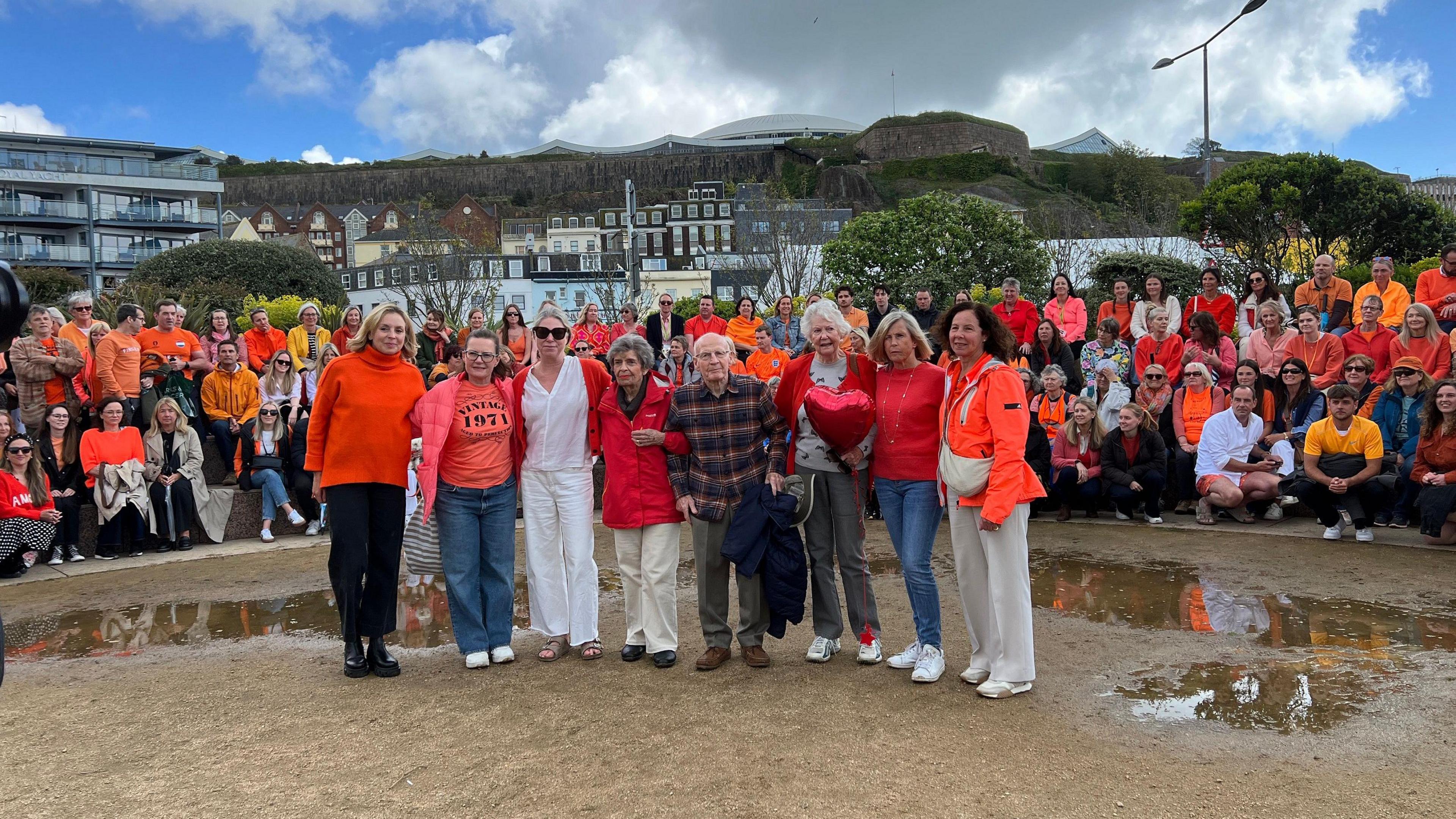 Hundreds of people are wearing orange and sitting on concrete steps. In front of them - Lily's family and close friends link arms - they're also wearing orange.
