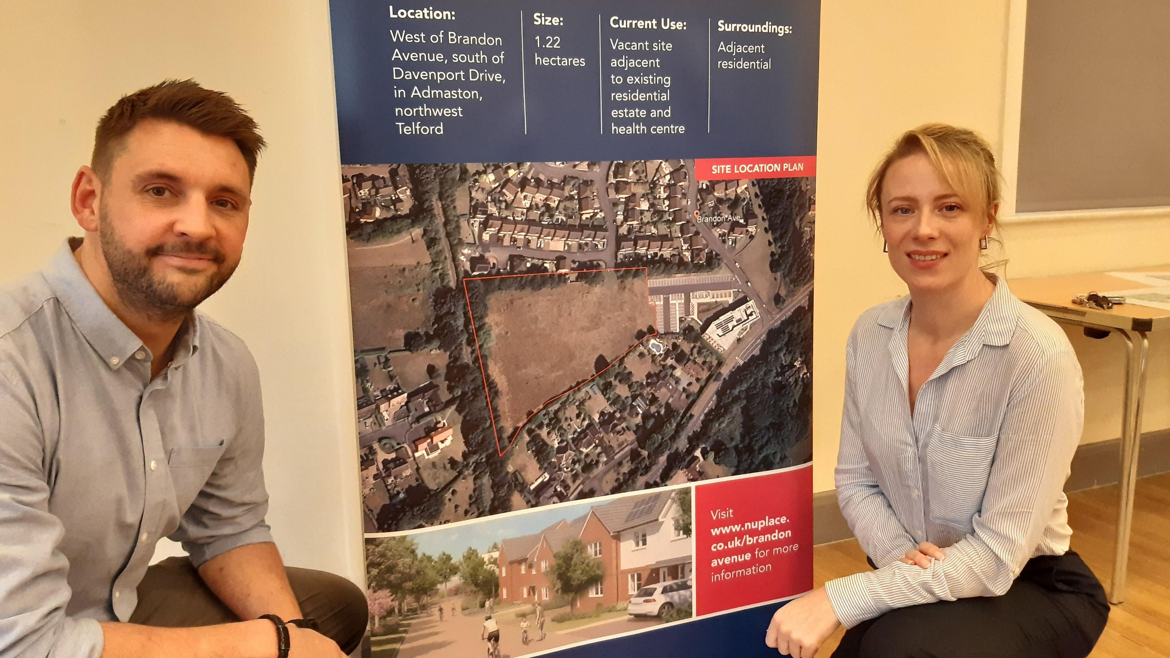 A man and a woman sit either side of a pop-up banner promoting a new housing development. The man has short brown hair and a cropped beard and is in an open-necked grey shirt, the woman has tied-back blonde hair and is wearing a patterned white blouse. They are in a meeting hall with a table behind them. 