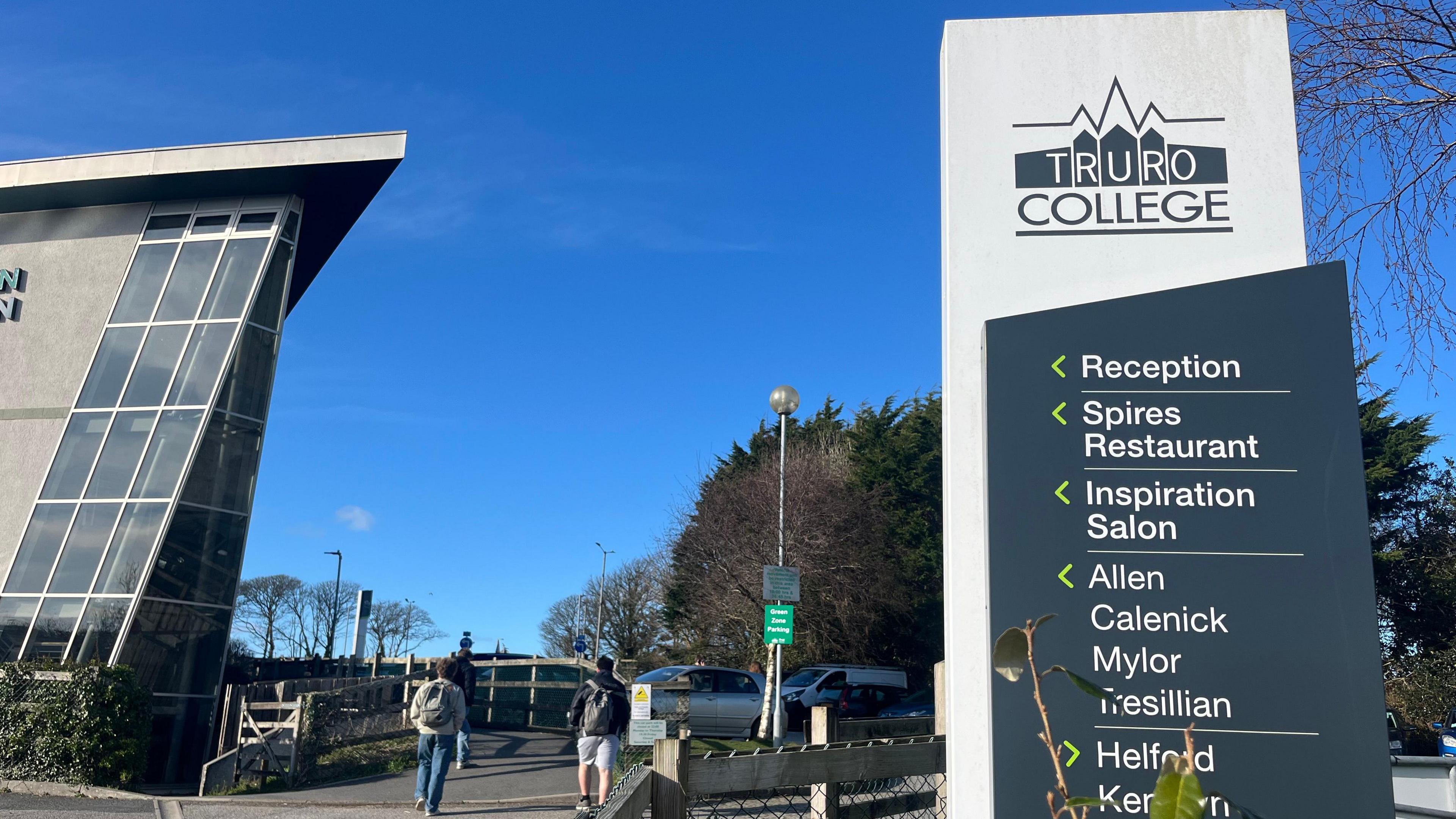 The entrance to the college. There is a tall sign that reads Truro College and arrows pointing to Reception, Spires Restaurant, Inspiration Salon, Allen, Calenick, Mylor, Tresillian and Helford. Two people with back packs are walking towards cars parked opposite a a glass fronted building. 