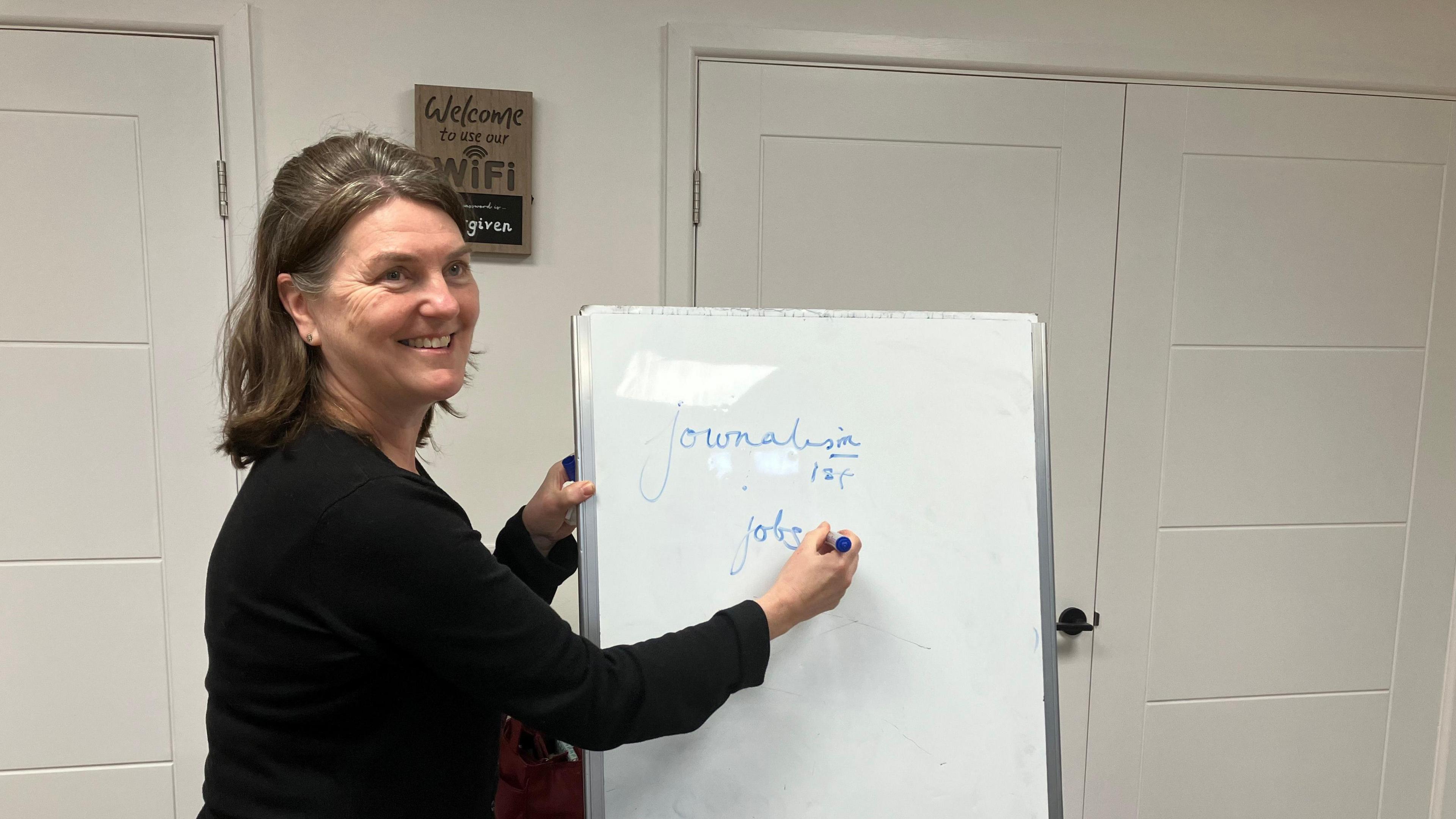 A picture of a woman with black hair and a dark grey skirt. She is stood in front of a white board with the words "journalism" and "jobs" on it.