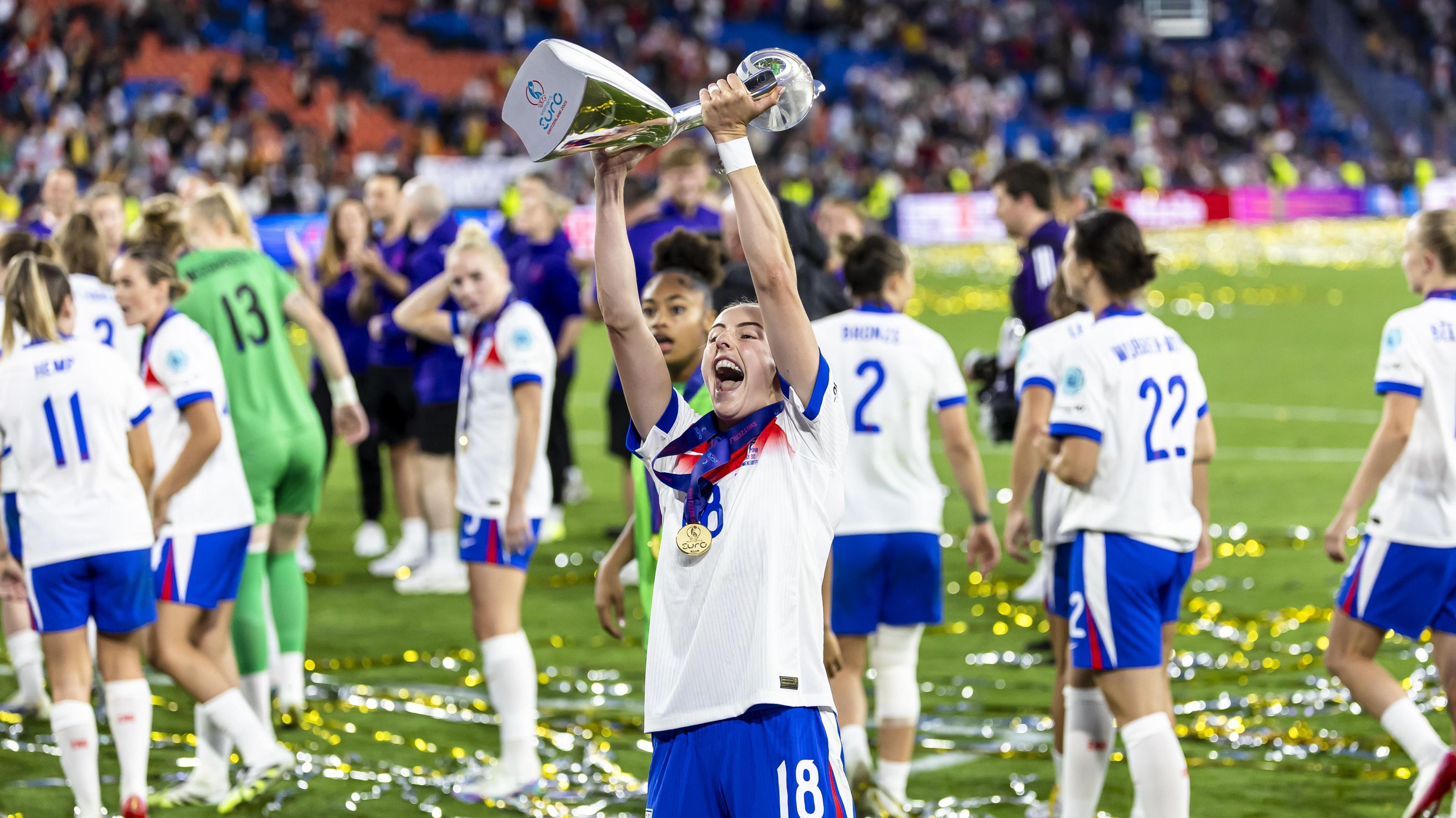 Chloe Kelly celebrates with the Euro 2025 trophy on the pitch with her team in the background.