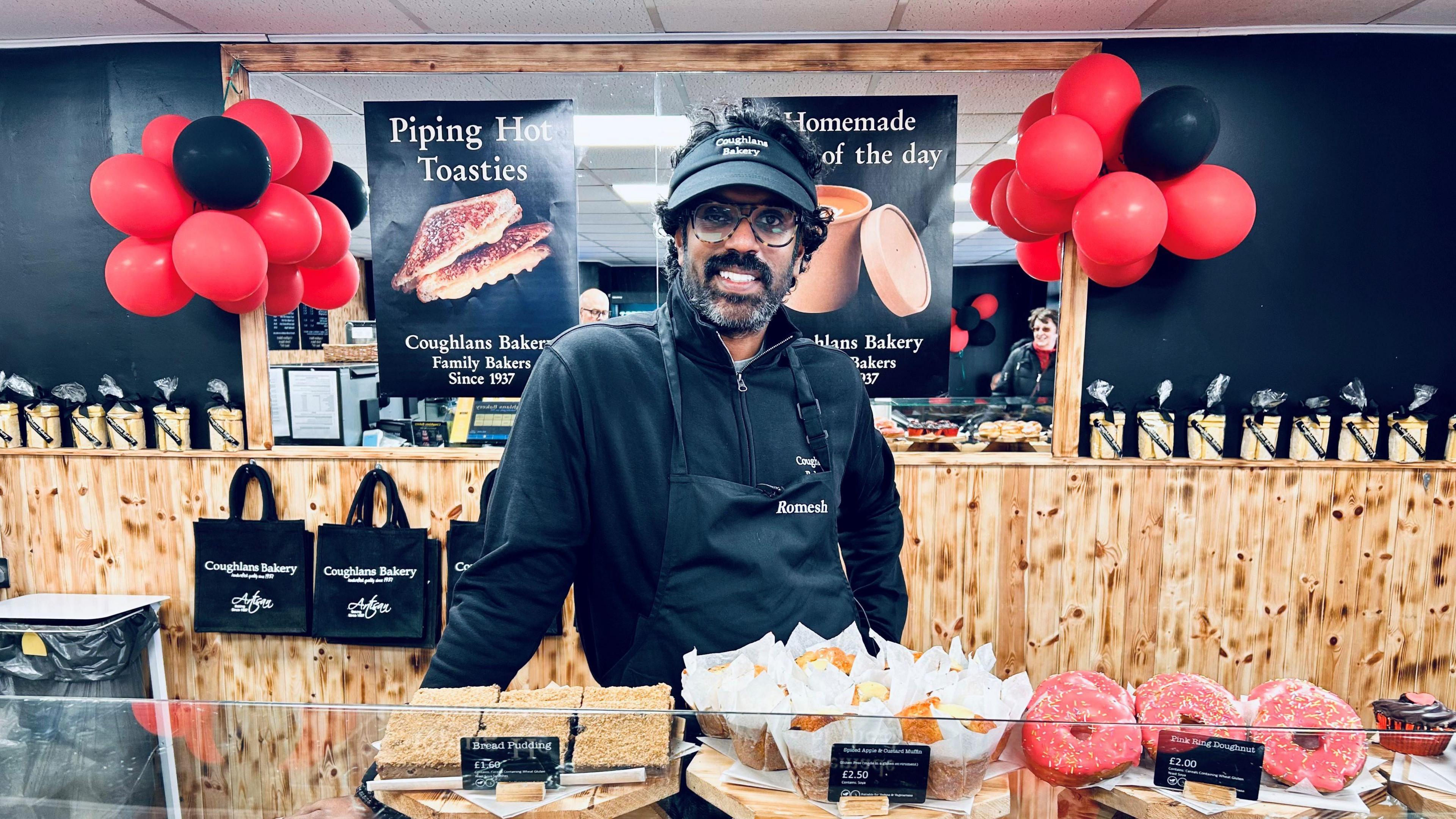 Romesh stood behind a counter of baked goods. He is wearing an apron and black cap and is smiling directly at the camera. There are red and black balloons behind him