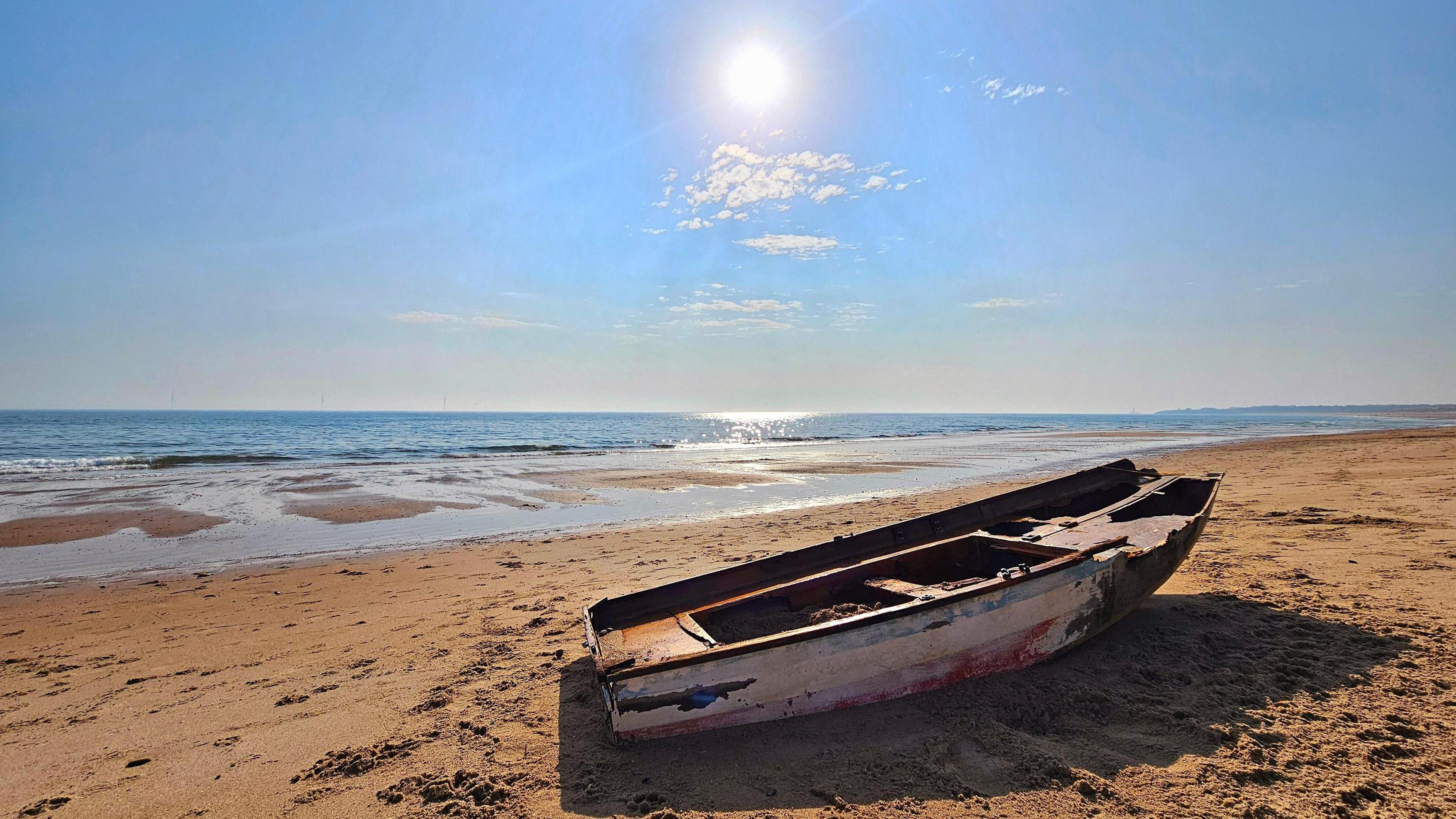 An abandoned wooden boat, with chipped paint, is on the sandy beach. The blue ocean is in the background and the bright sun is casting shadows.