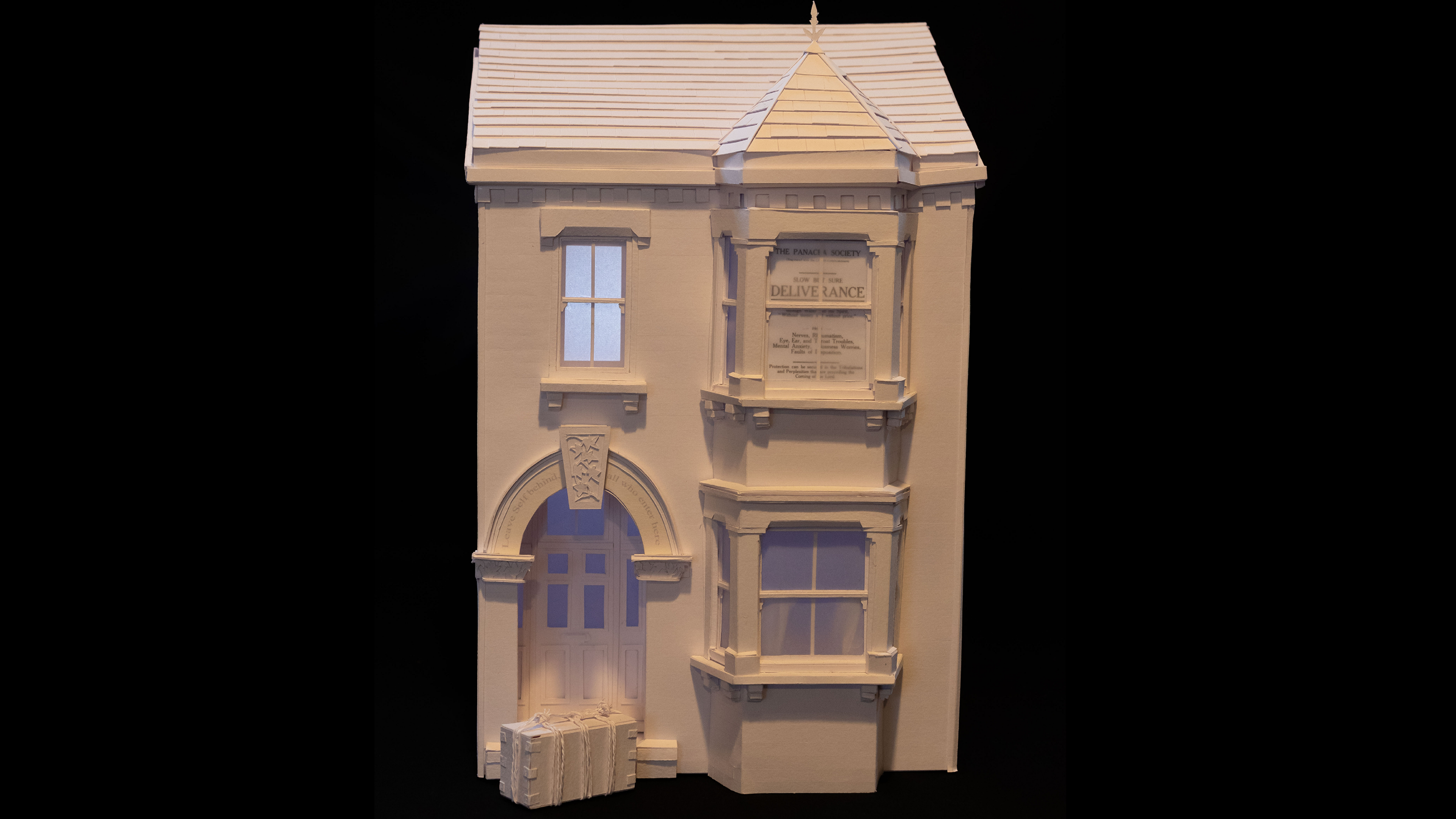 A white paper sculpture of an Edwardian house, with a box outside the front door, it has three windows, a front door, and a slate roof. A light is on inside and the rest of the image is in black. 