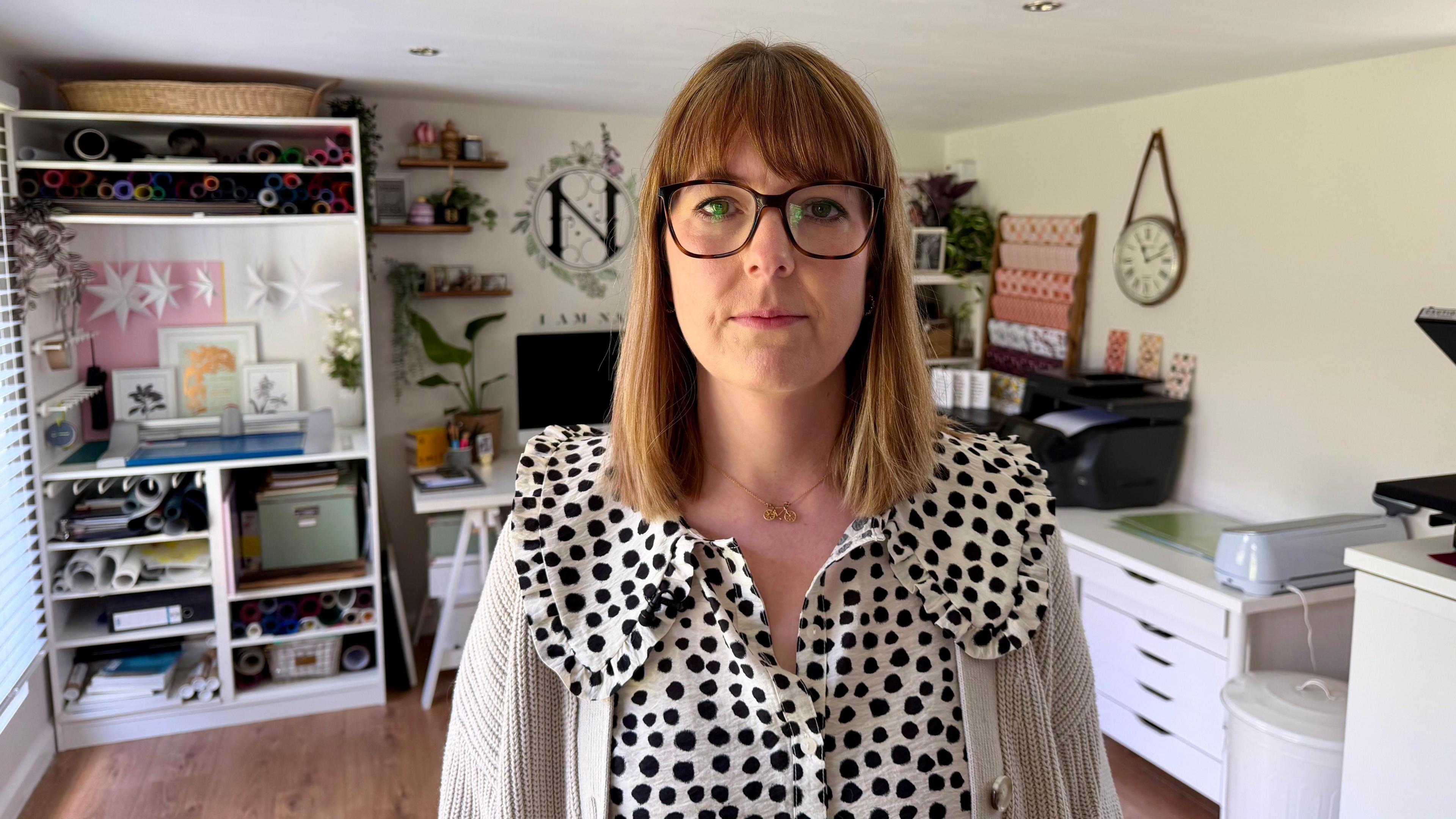 Natalie Ramsell-Lee. She has short brown hair, a fringe and wears glasses. She is also wearing a black and white polkadot blouse. She is sat her desk, with one of her designs on the screen, and is smiling at the camera.