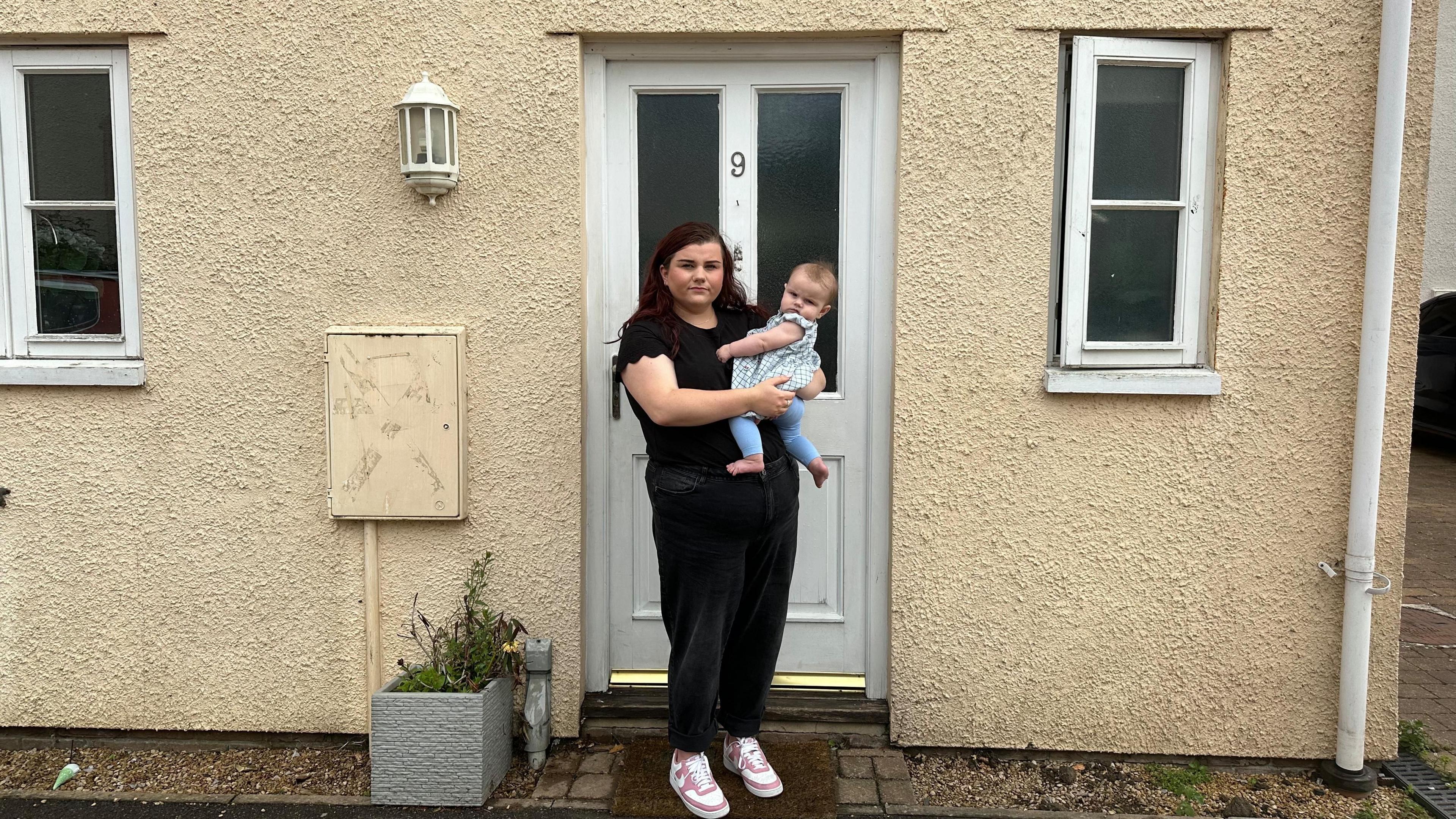a woman dressed in black holding her baby on her hip standing outside the white front door of a terraced cream-painted house