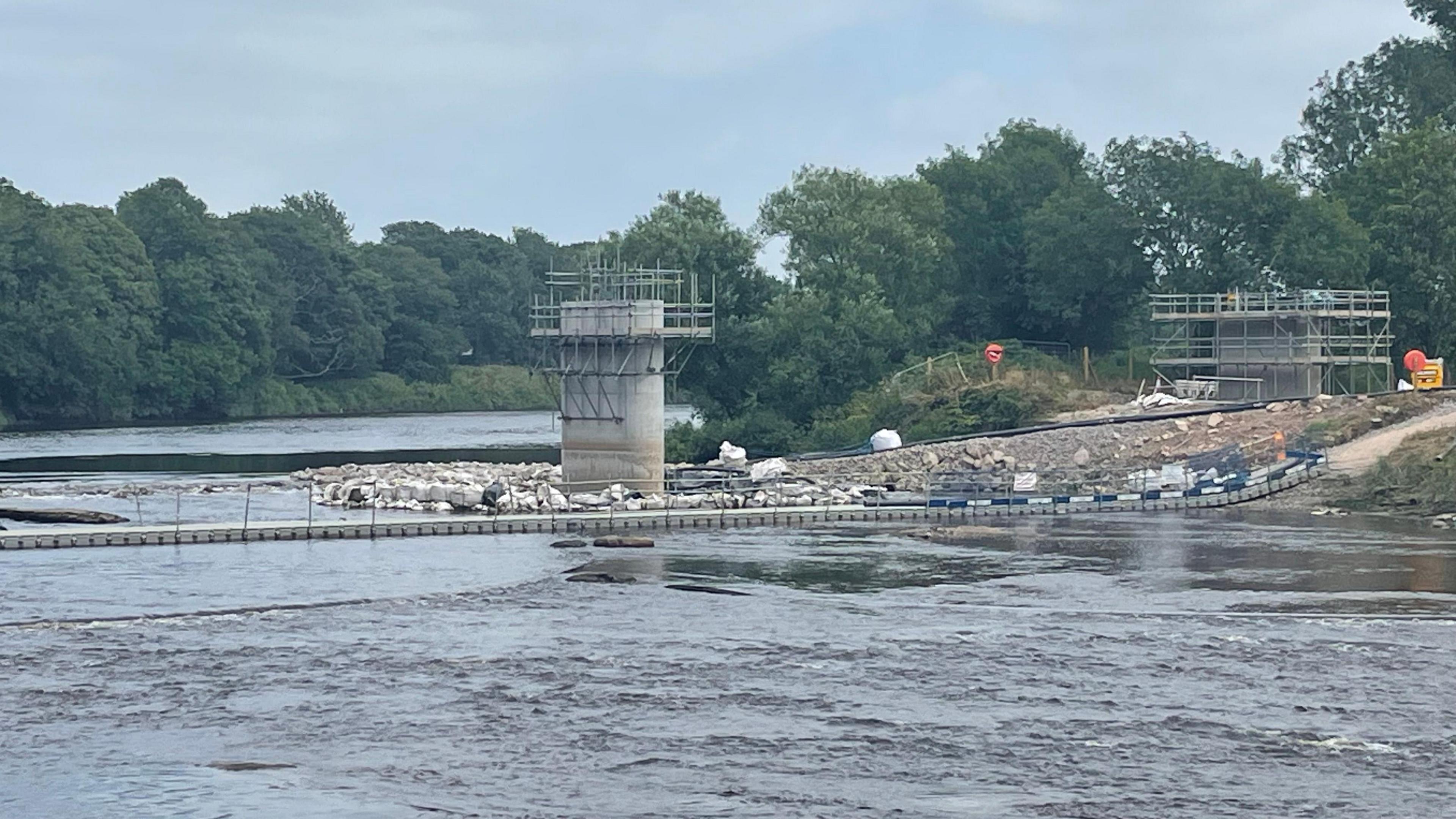 An image of the River Ribble where work is being carried out to build a new bridge. To the left, there are two concrete pillars with scaffolding around them, with rubble and dirt piles. In the river there are bollards made of concrete along the line of the new bridge. There are trees in the background.