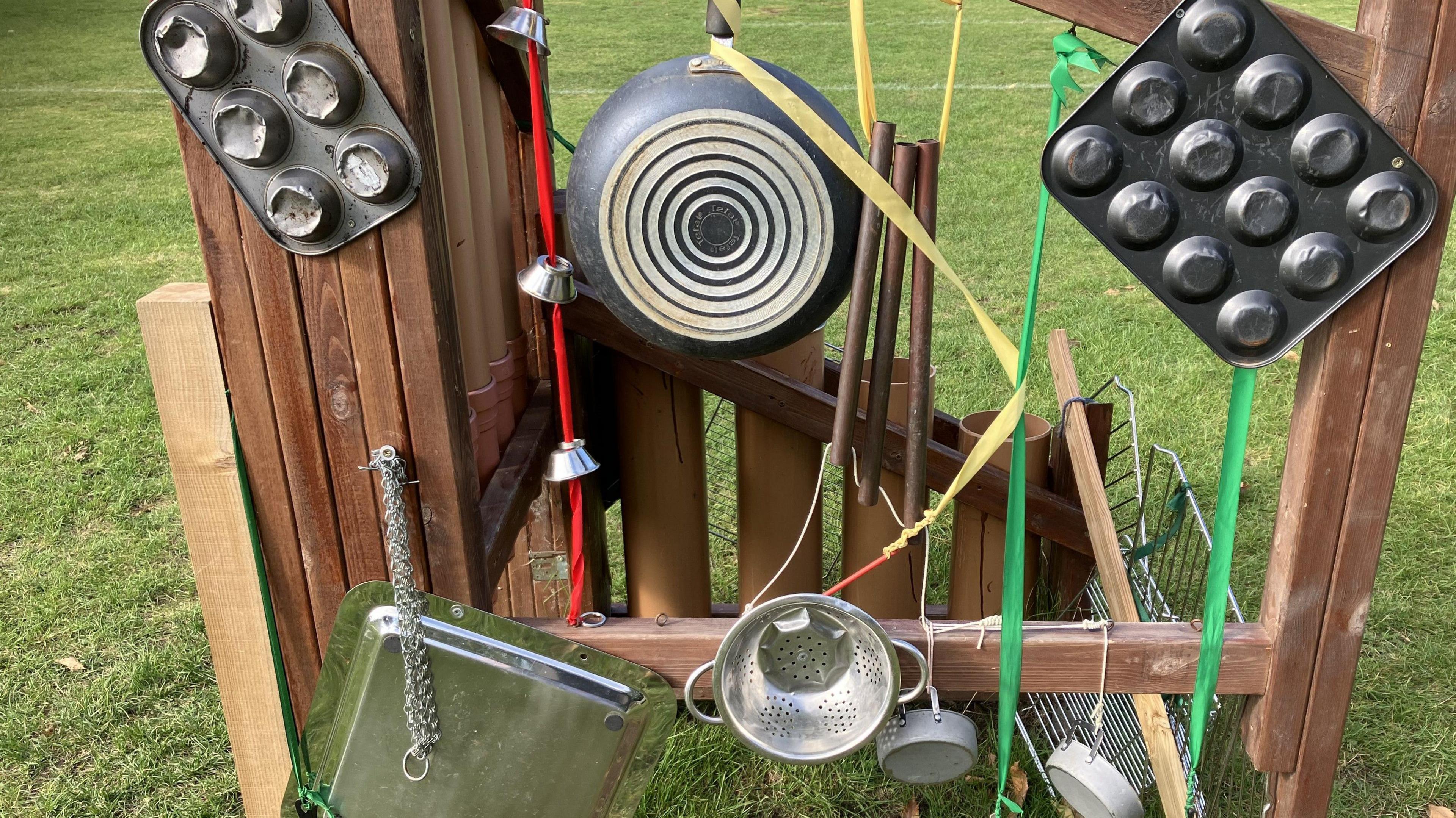 An array of kitchen utensils and frying pans are hanging from a wooden frame in playground where children can create sounds and rhythms 