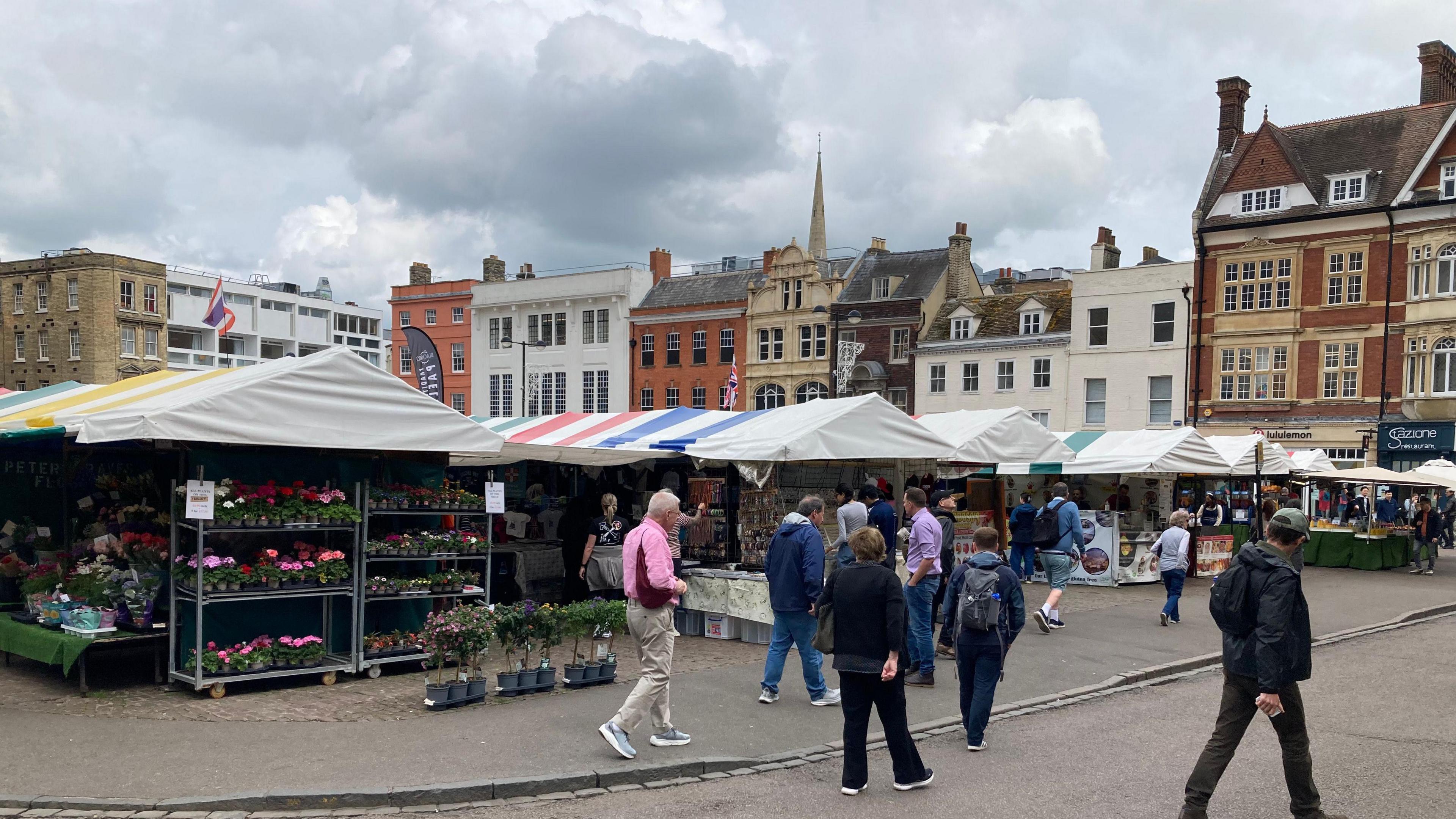 A wide shot of Cambridge Market Square. At the front is a plant stall and a number of people are walking by. Tall buildings are visible behind.