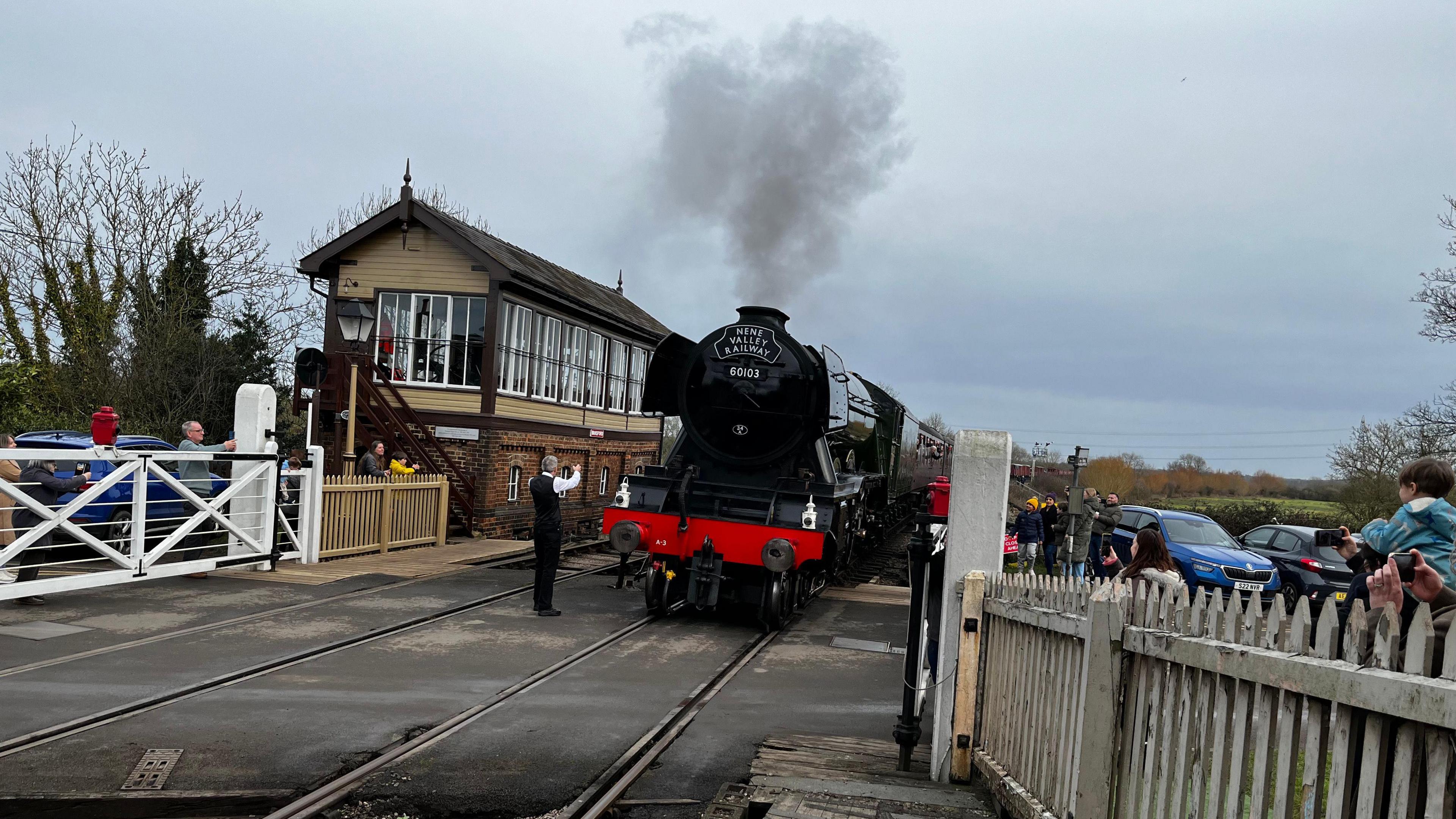 The steam engine approaches a level crossing. Crowds of people either side film on their phones. The loco has a plume of black smoke coming from its funnel. A smartly dressed man is stood in front, guiding it in with arm gestures.