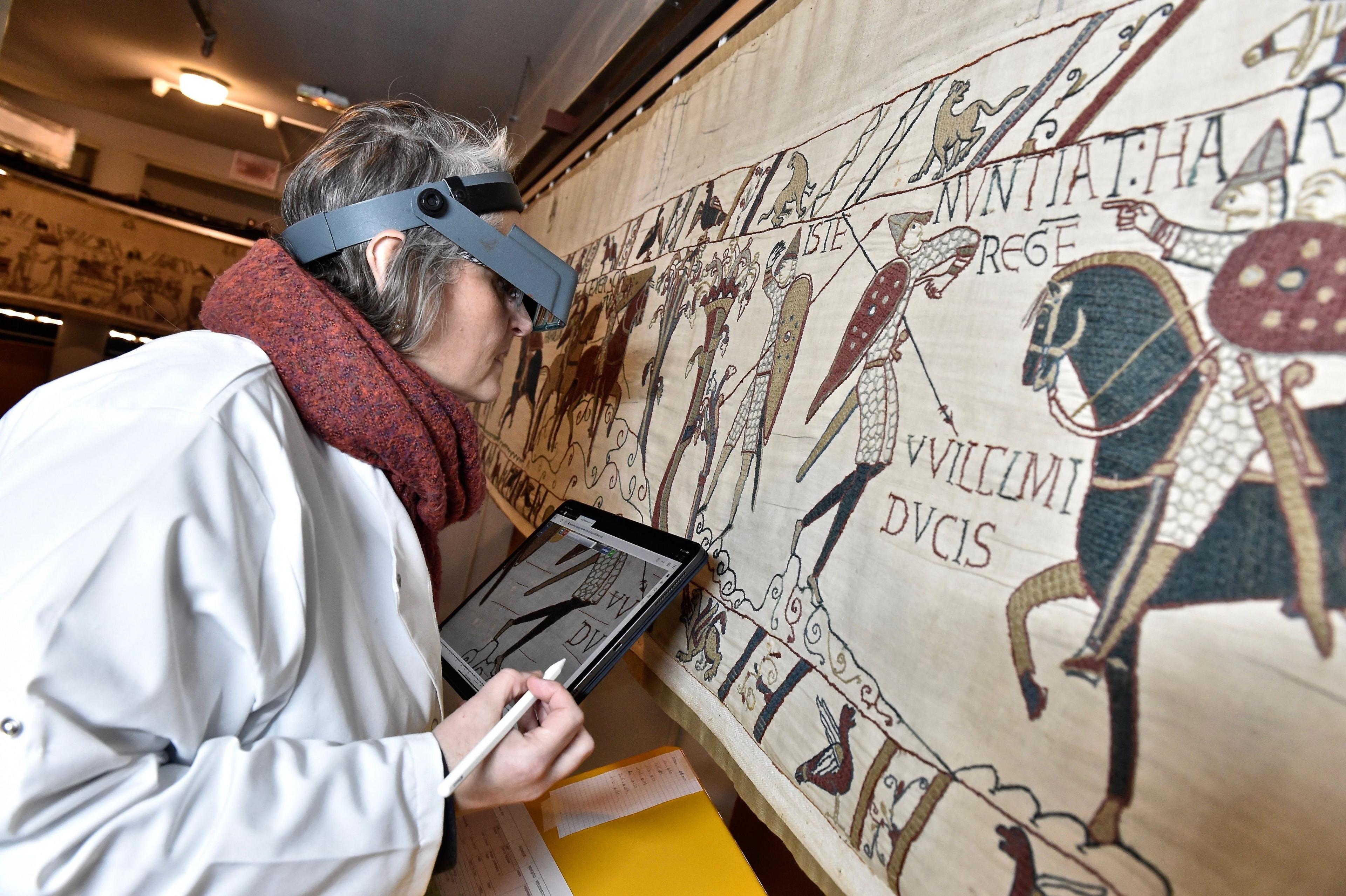 A woman with a white coat, visor and iPad looks closely at a scene from the Bayeux Tapestry