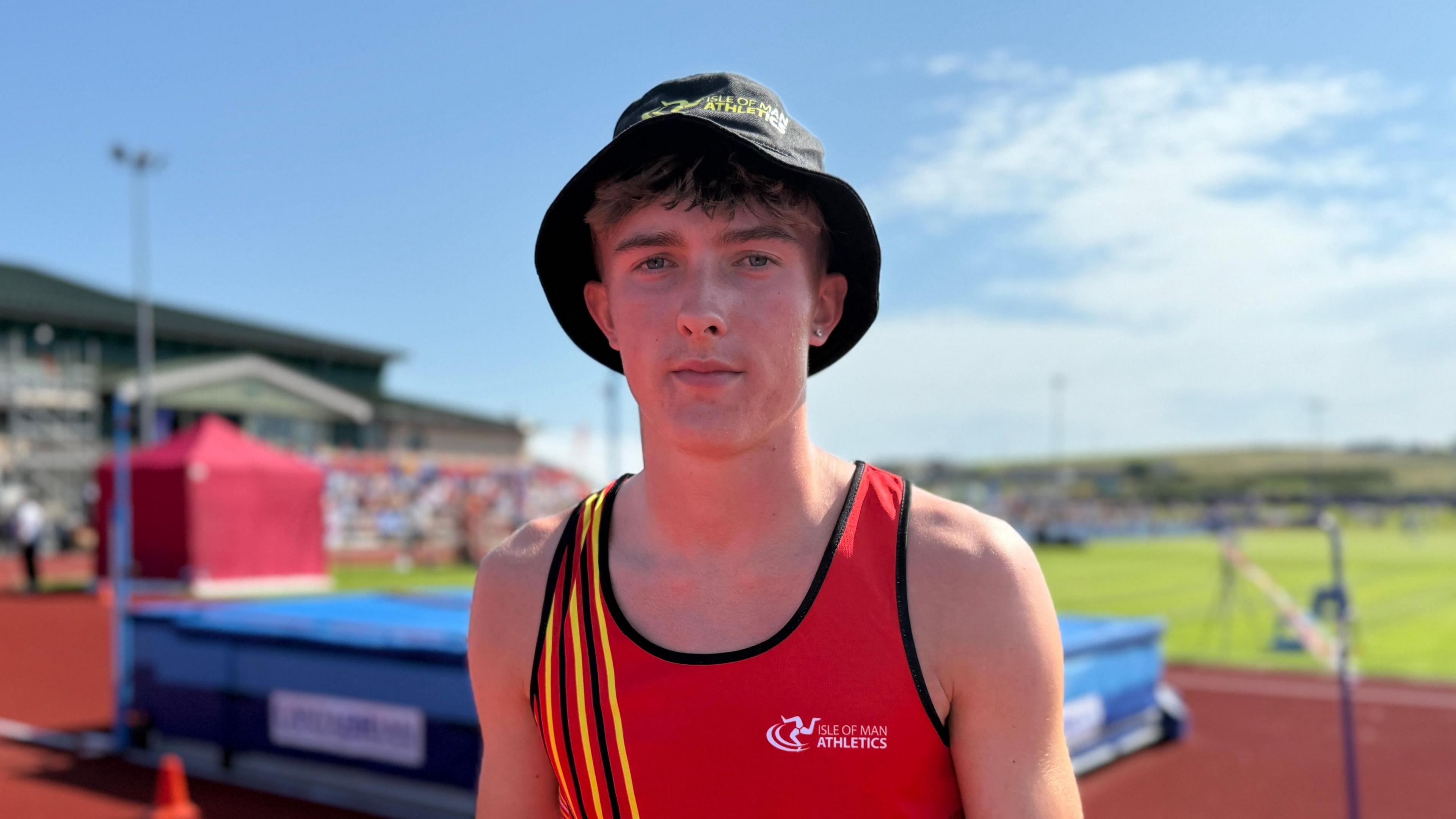 Regan Corrin wars a black bucket hat and a red vest. He stands near the high jump equipment of a bar and a blue crash mat by an orange race track.