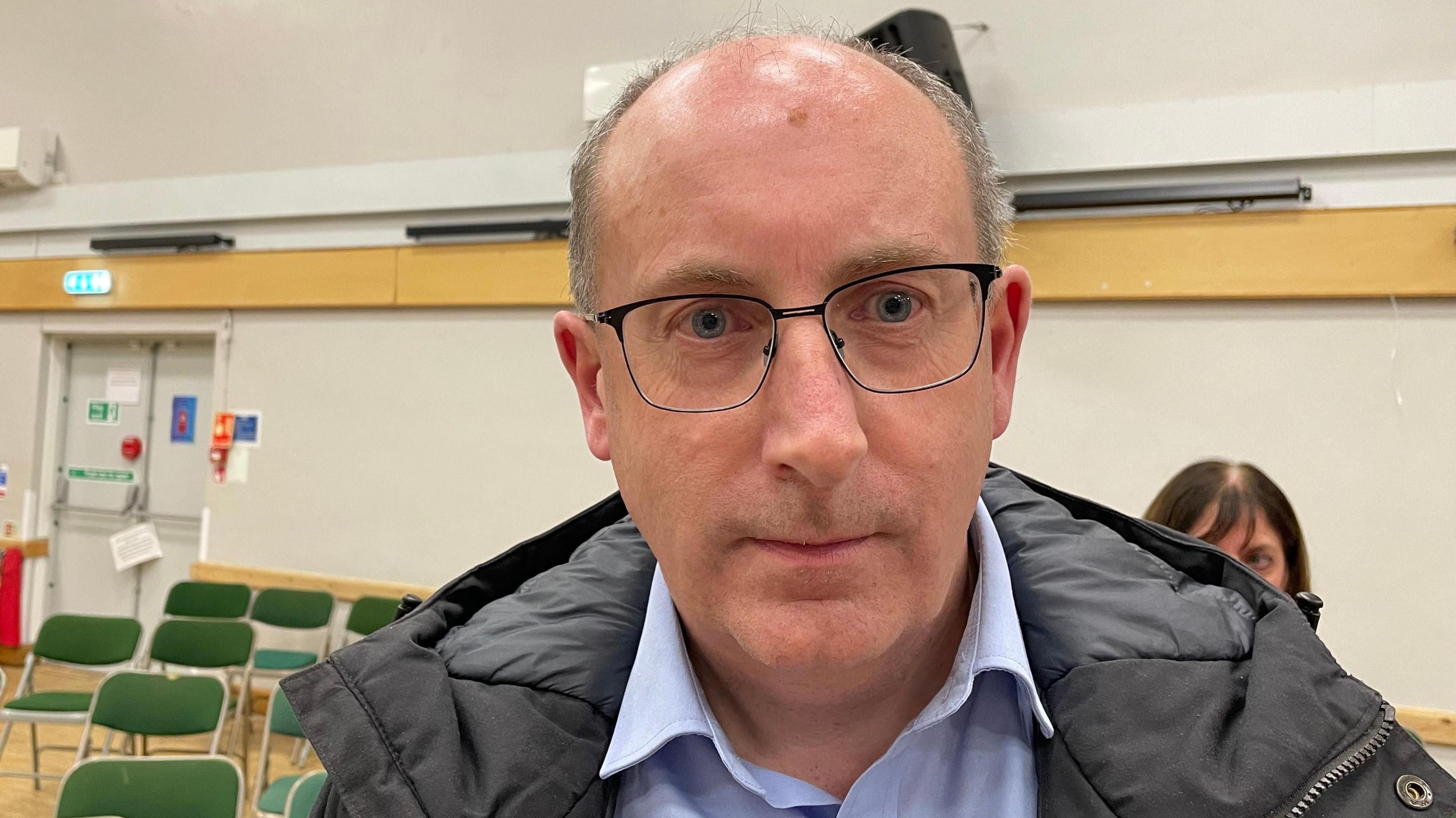 Nick Brown wearing a light blue collared shirt, black coat and glasses.