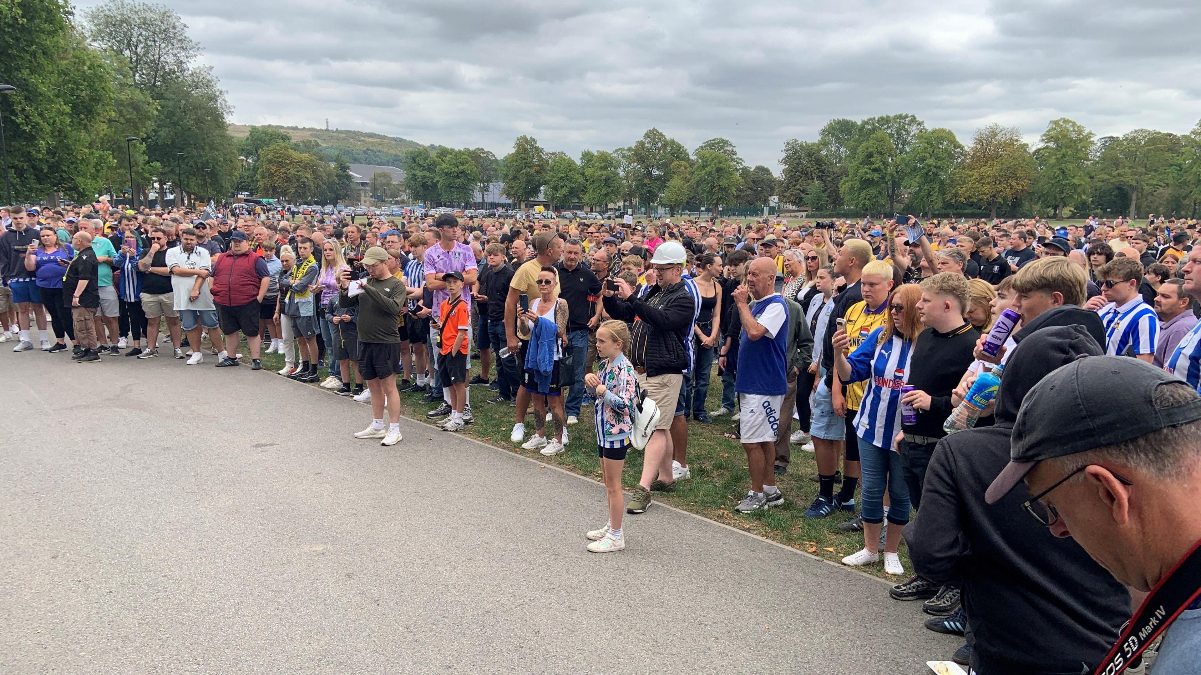 Hundreds of football fans, some in SWFC shirts, are gathered in a park - they form a large crowd and some are taking photos of the protest. 
