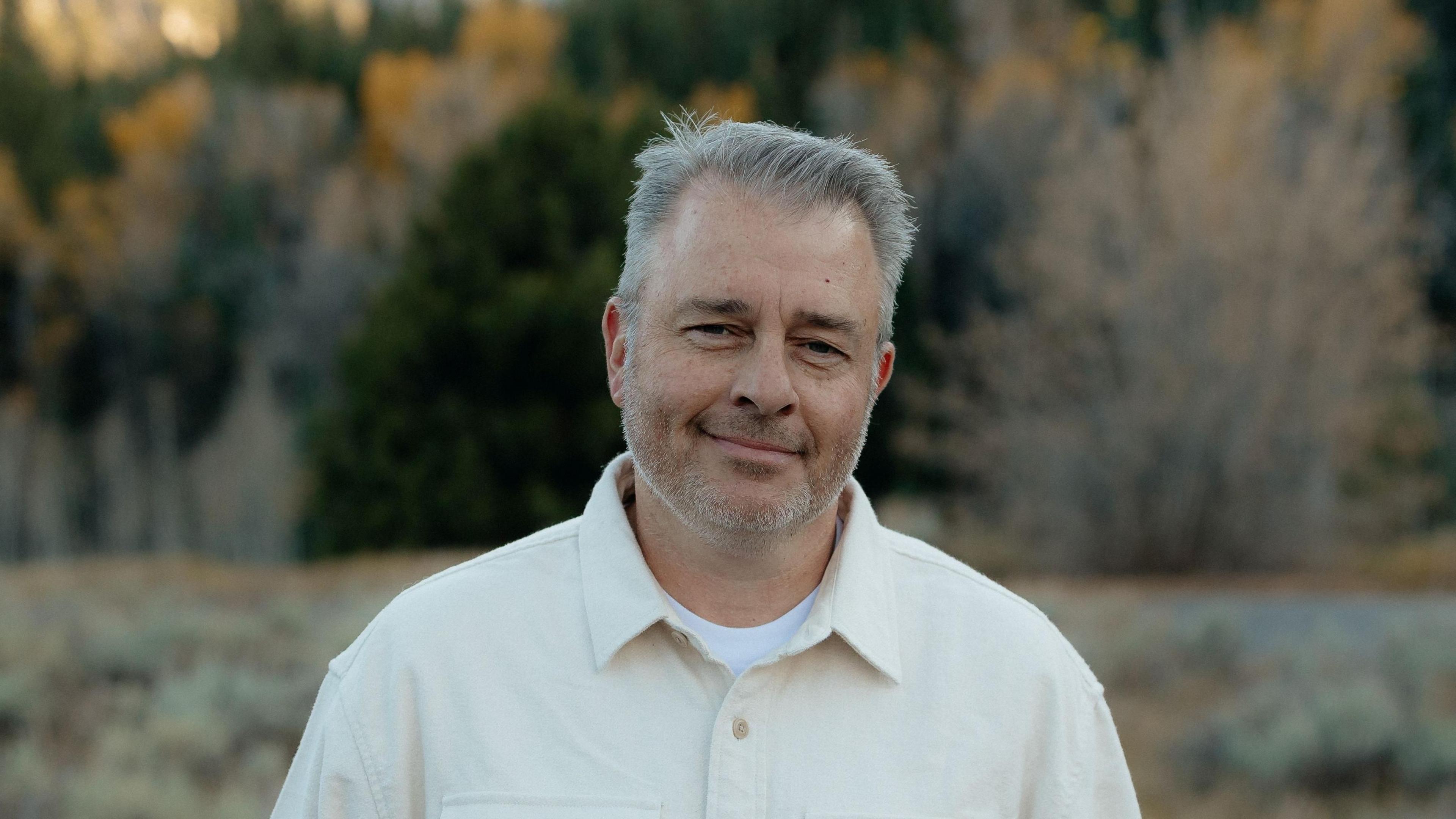 Jared Hendricks, in pale polo shirt with white undershirt, stands in front of field and trees