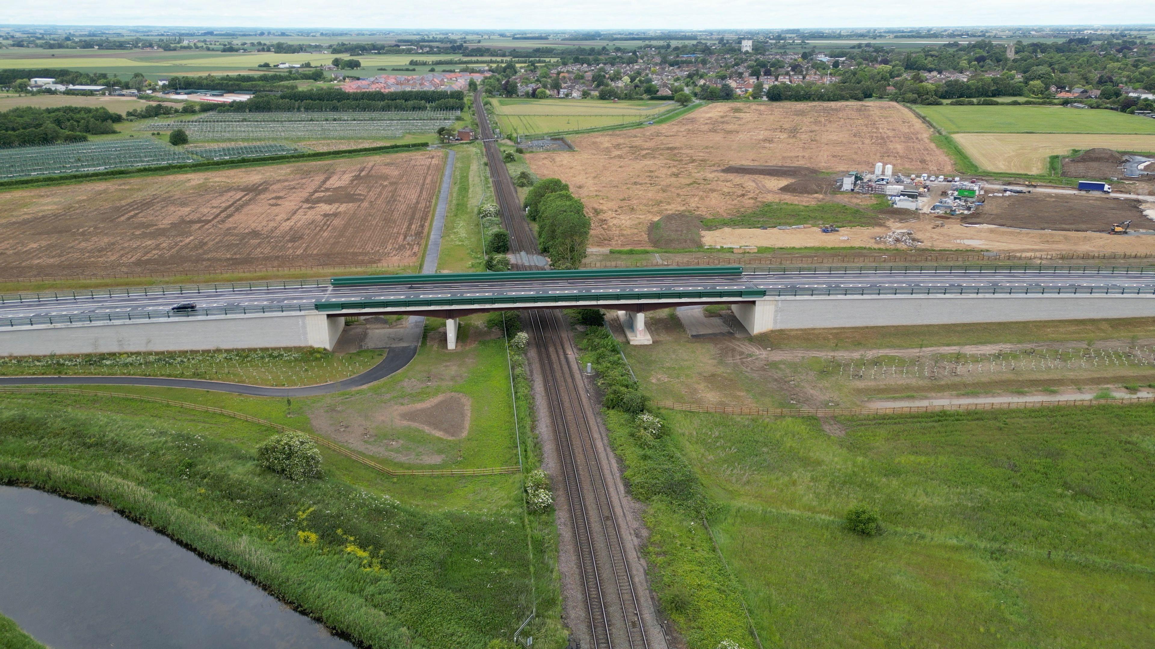 The newly-built grey bridge runs right to left above a railway line which passes underneath it. The village of Pinchbeck is in the distance but green fields surround the bridge and railway line.