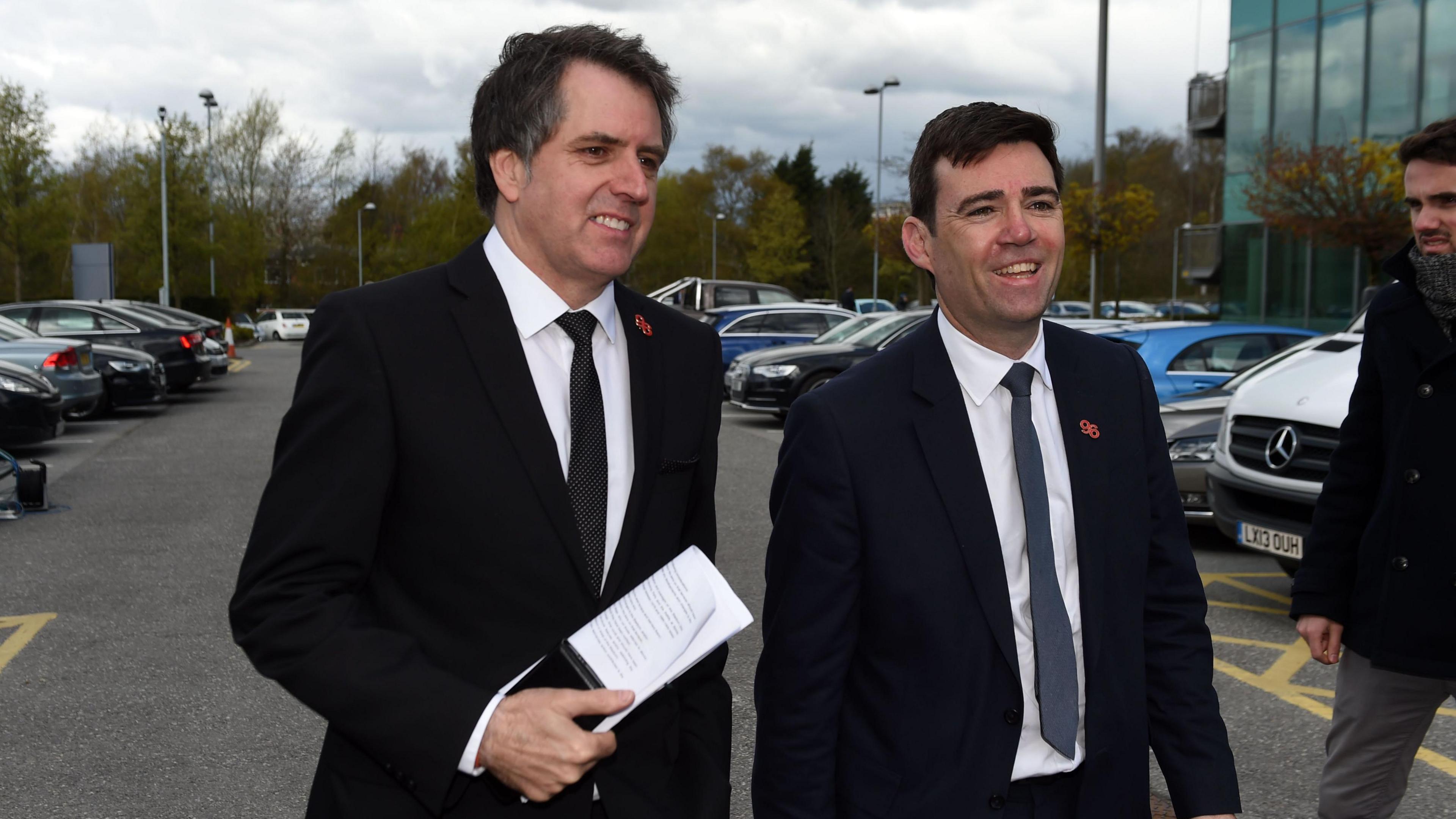 Two white middle aged men with short hair wearing dark suits, white shirts and ties walk through a car park, smiling broadly. 