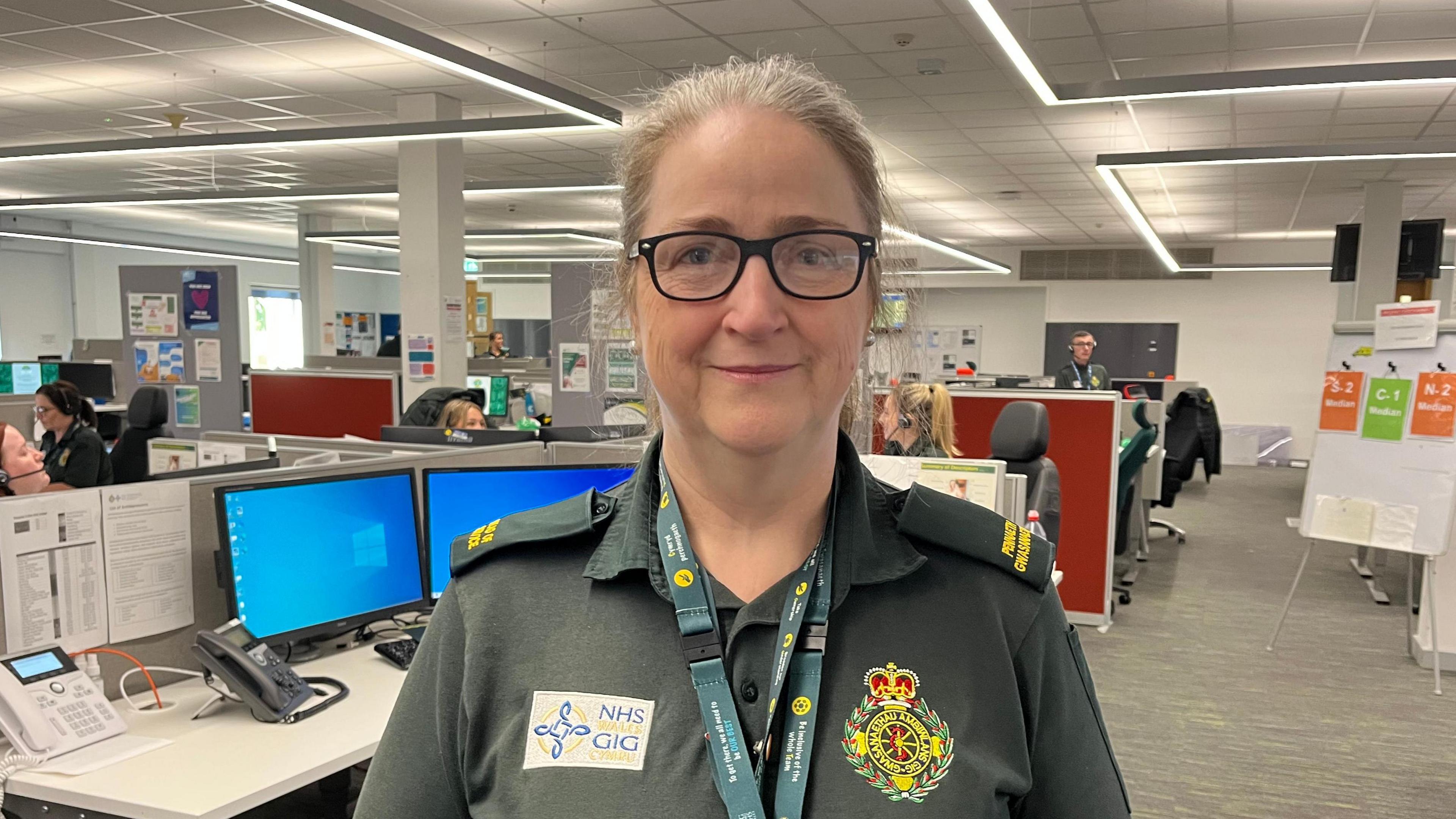 Ambulance service worker Gill Pleming, wearing a dark green ambulance service polo shirt carrying the NHS Wales logo, smiles at the camera. Behind her, staff in the ambulance control room wear headsets and work at their screens. It is a head and shoulders shot of her.