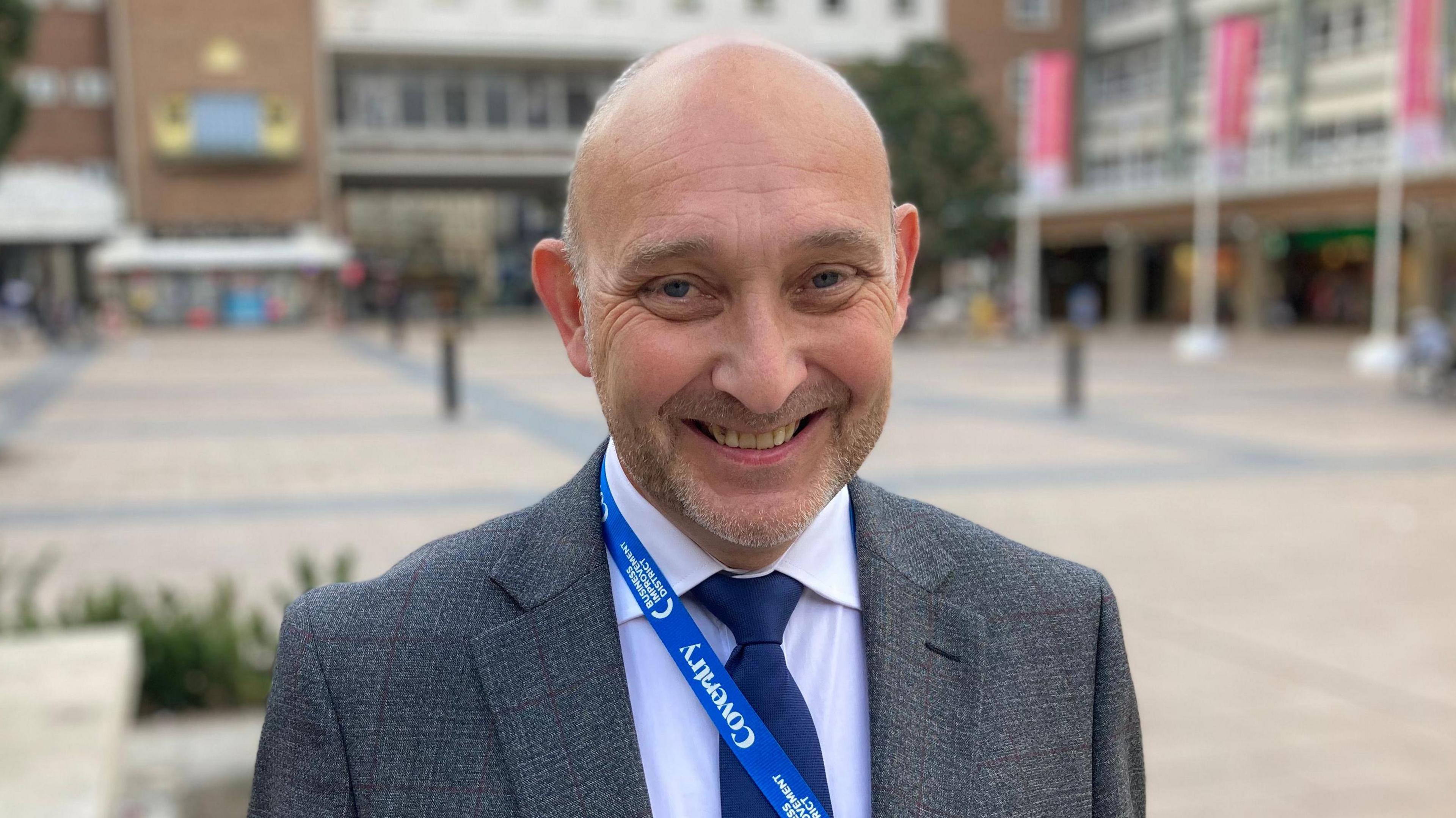 A man wearing a grey suit , blue tie and white shirt smiles toward the camera, behind him are buildings and a public square.