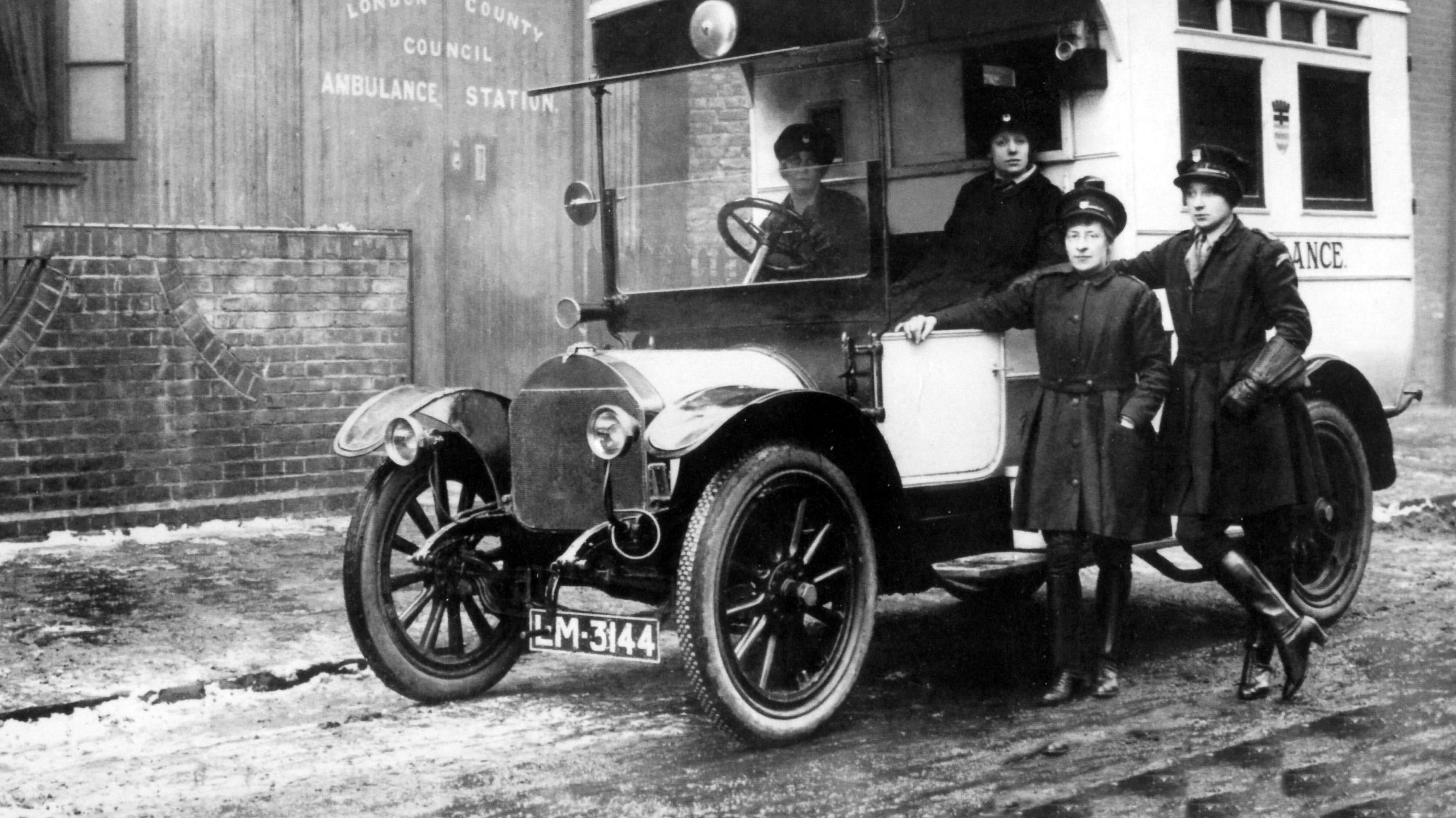 Two women in an old ambulance look at the camera while two women stand outside it looking at the camera, in uniform