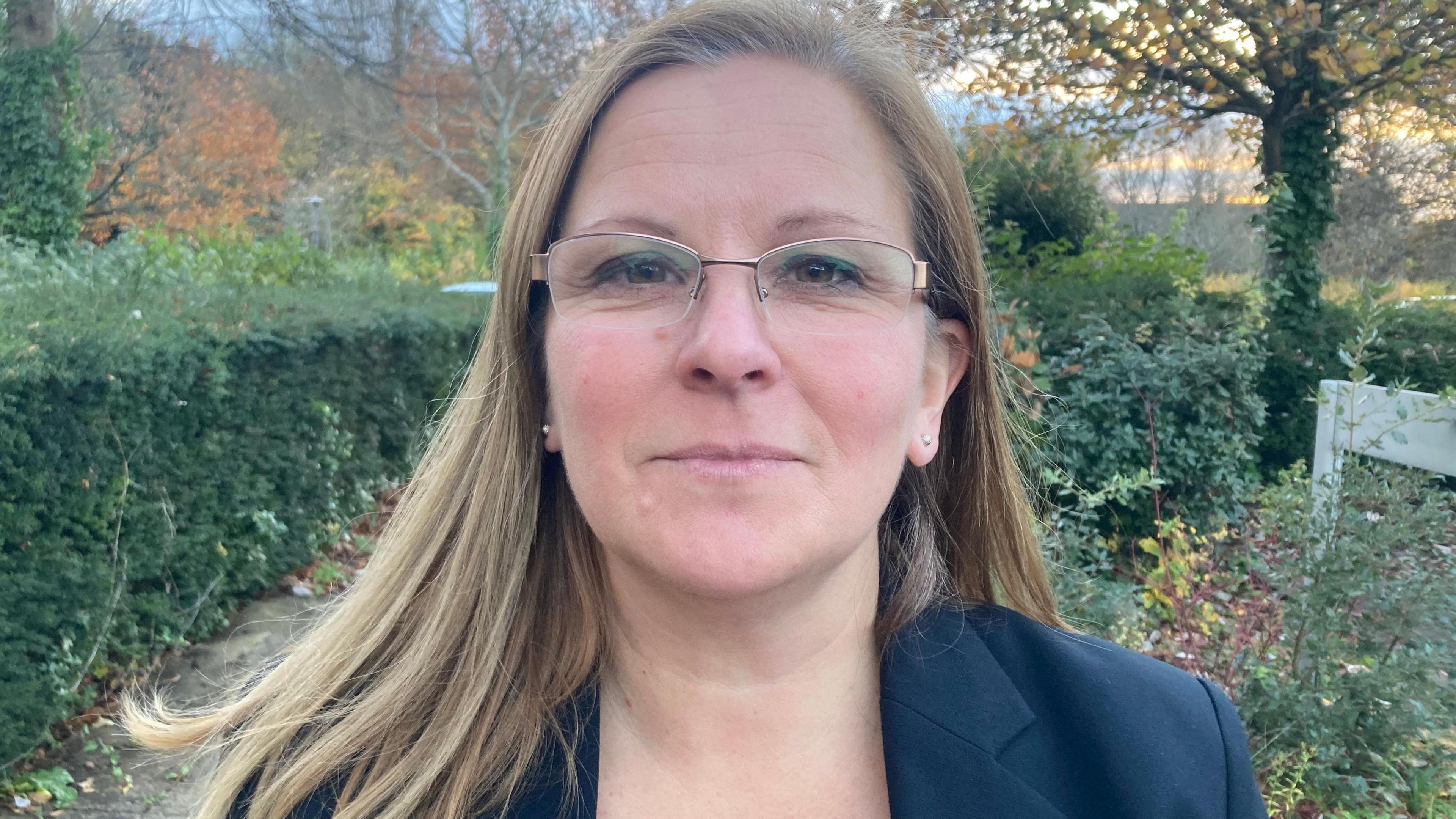 A woman wearing glasses looking at the camera, wearing a navy blazer