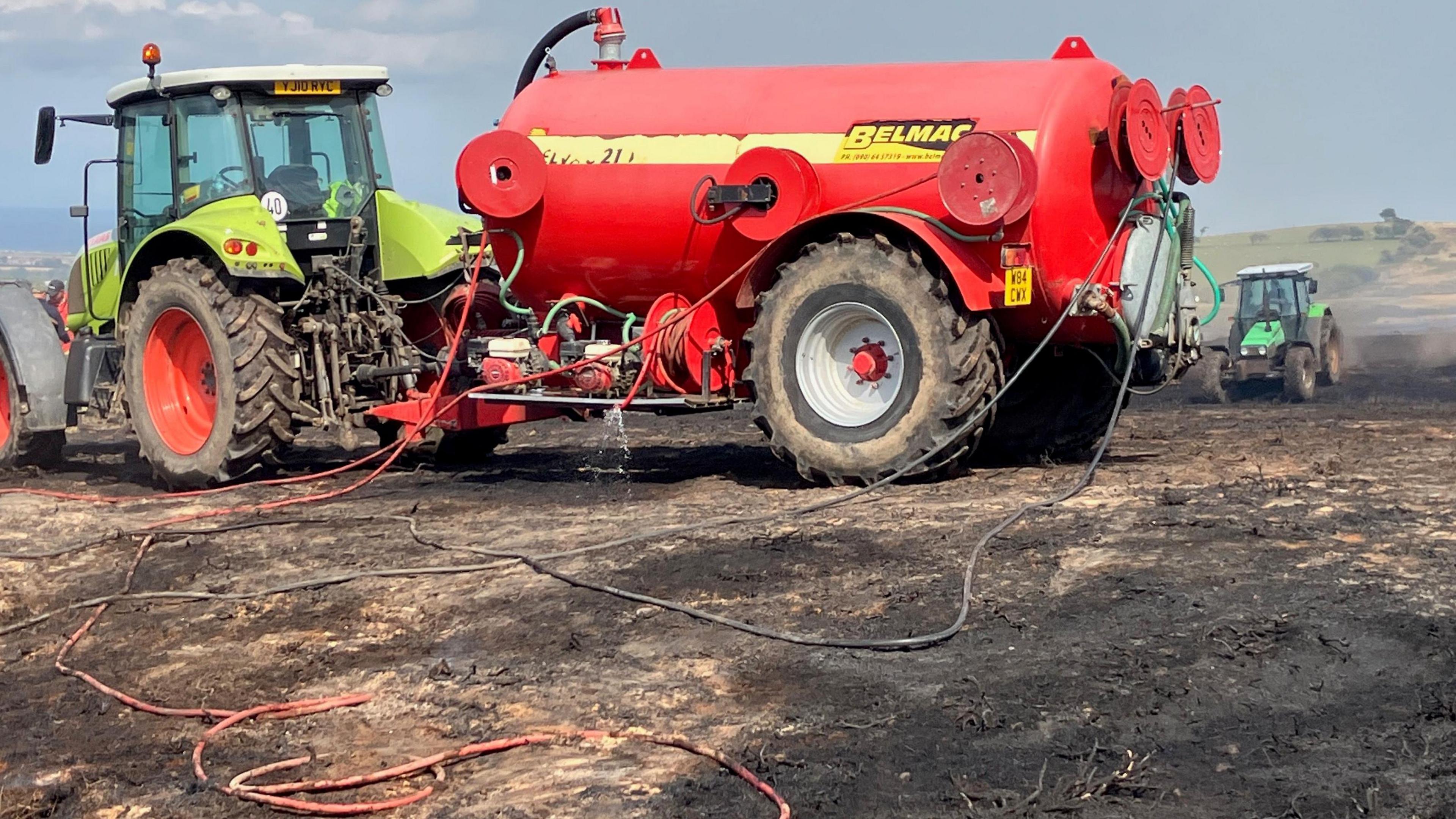 A tractor pulling a red slurry tank containing water across burnt out moorland witha nother tractor seen in the distance